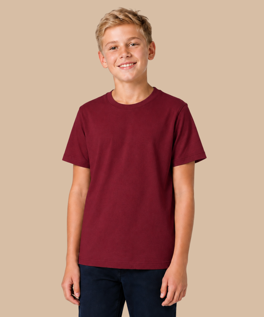 A smiling young male model wearing a solid maroon crewneck t-shirt and dark blue jeans, standing against a solid beige background. Handprinted t-shirt services from Pathfinder Printworks.