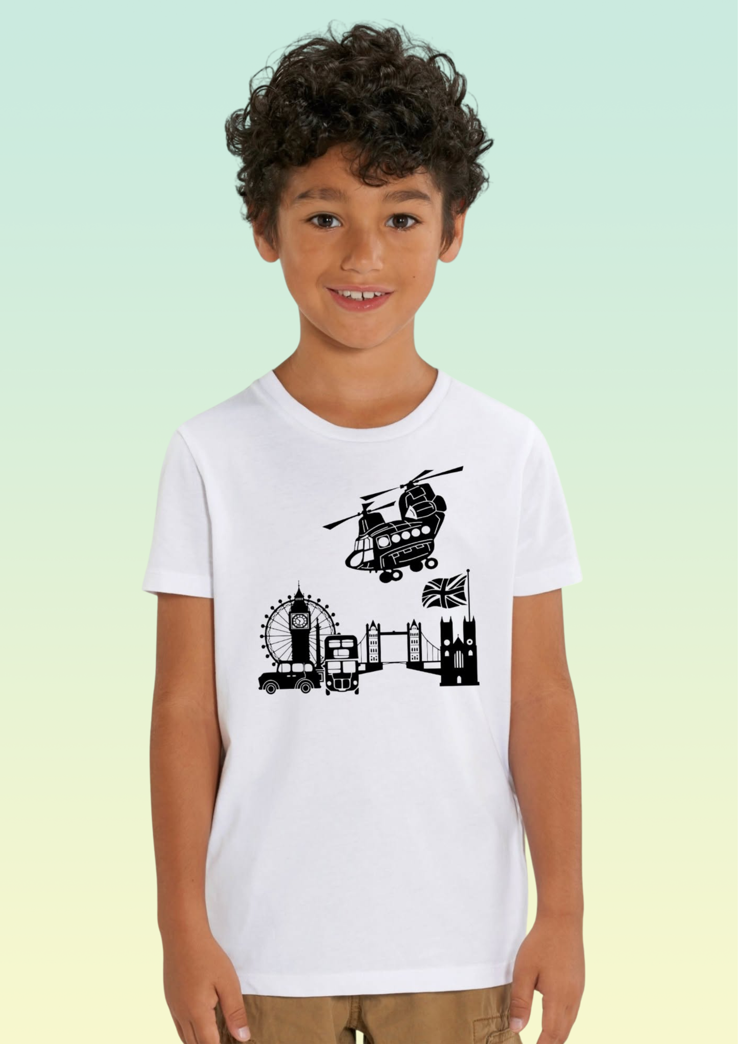 A young boy with curly dark hair wearing a white short-sleeve t-shirt with a black graphic print of London landmarks (Big Ben, London Eye, Tower Bridge, Westminster Abbey, and a helicopter) on the front, set against a light green and yellow gradient background. Exclusive to Flying Teez
