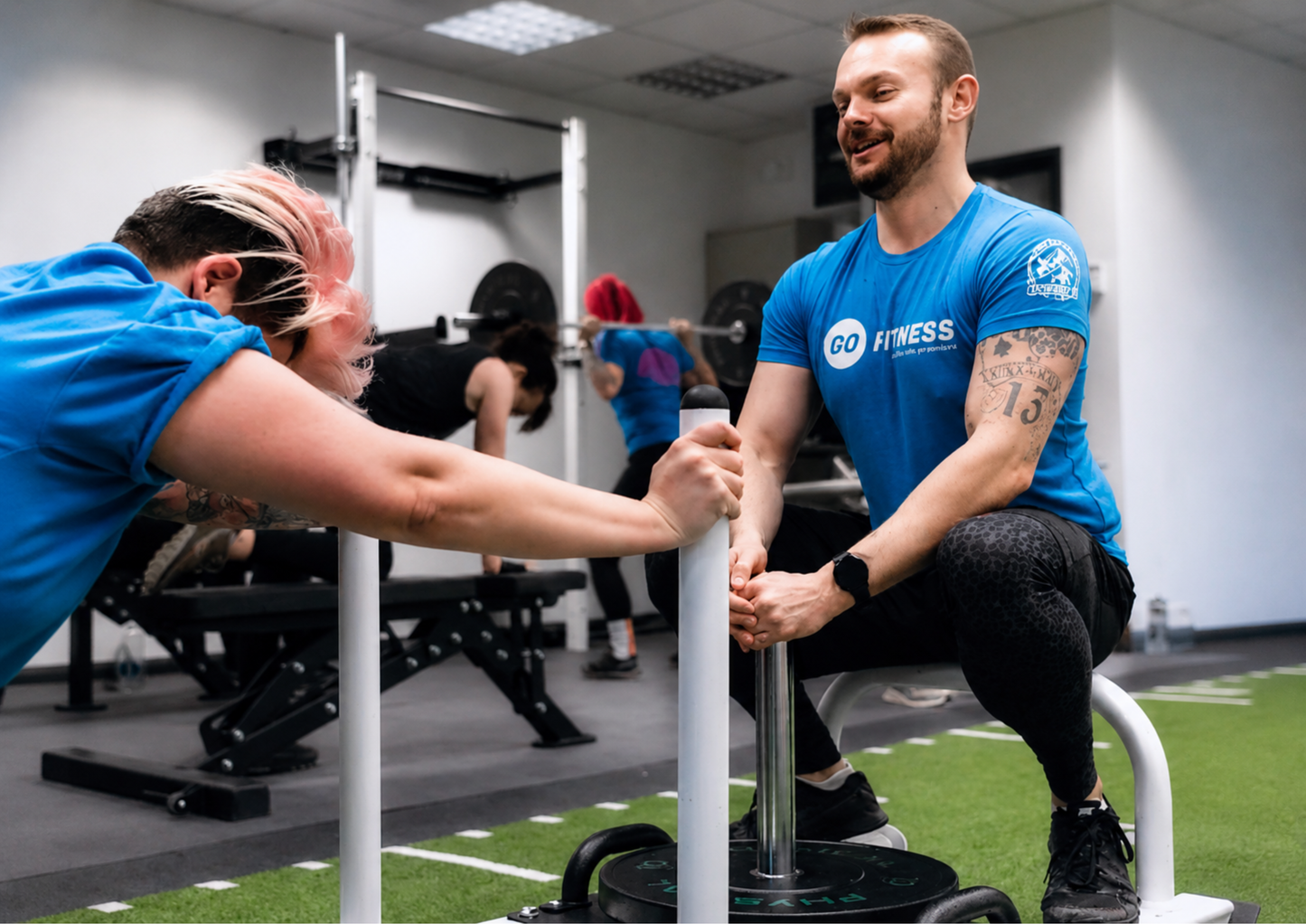 A personal trainer coaches a client during a gym session while wearing a branded fitness t-shirt. The image highlights community, coaching, support and professional teamwear in an active training environment.