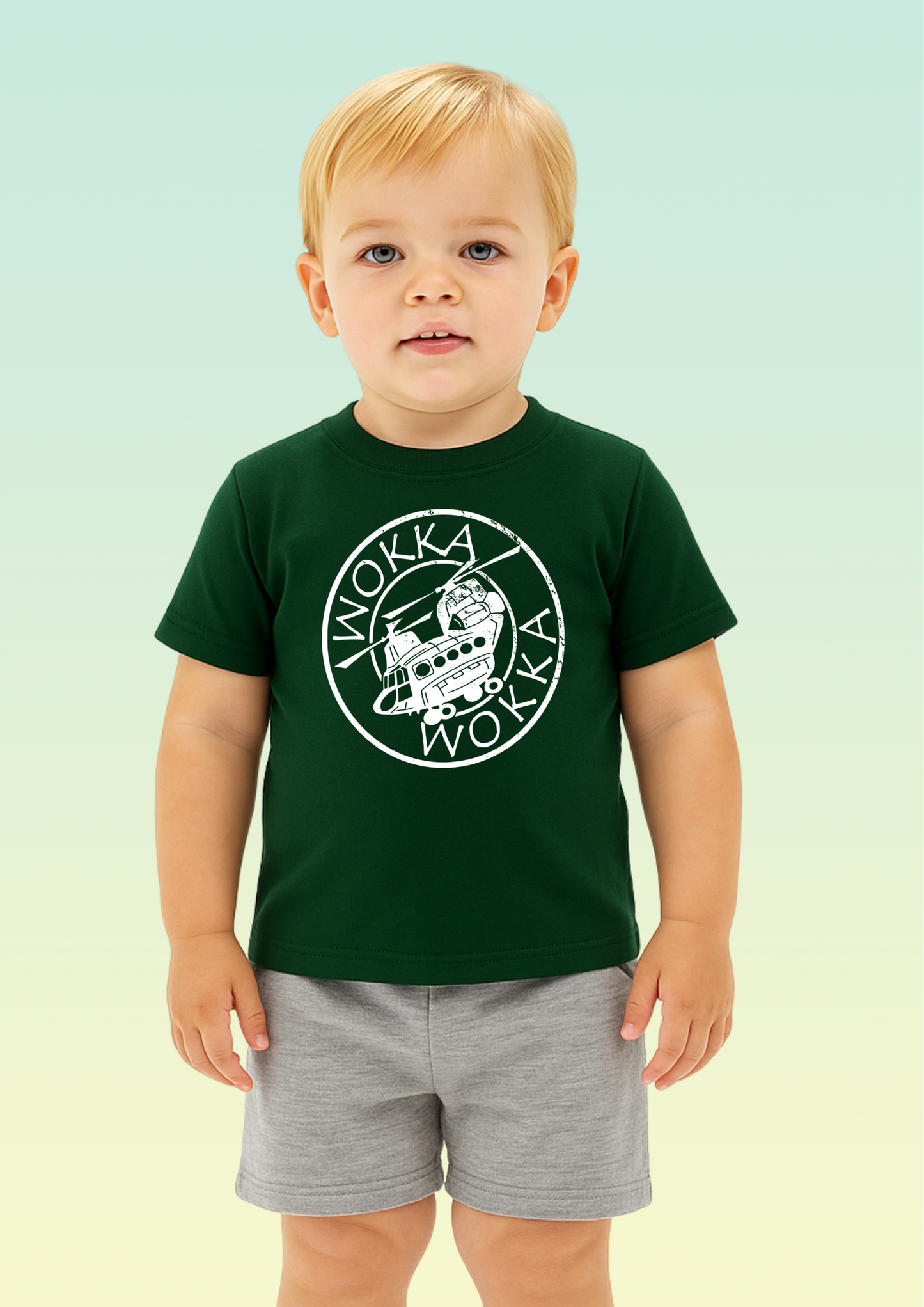 A young toddler with blond hair stands against a pale mint-to-yellow gradient background, wearing a dark green short-sleeve T-shirt with a large white circular 'Wokka Wokka' graphic on the chest and light gray shorts. Exclusive to Flying Teez.