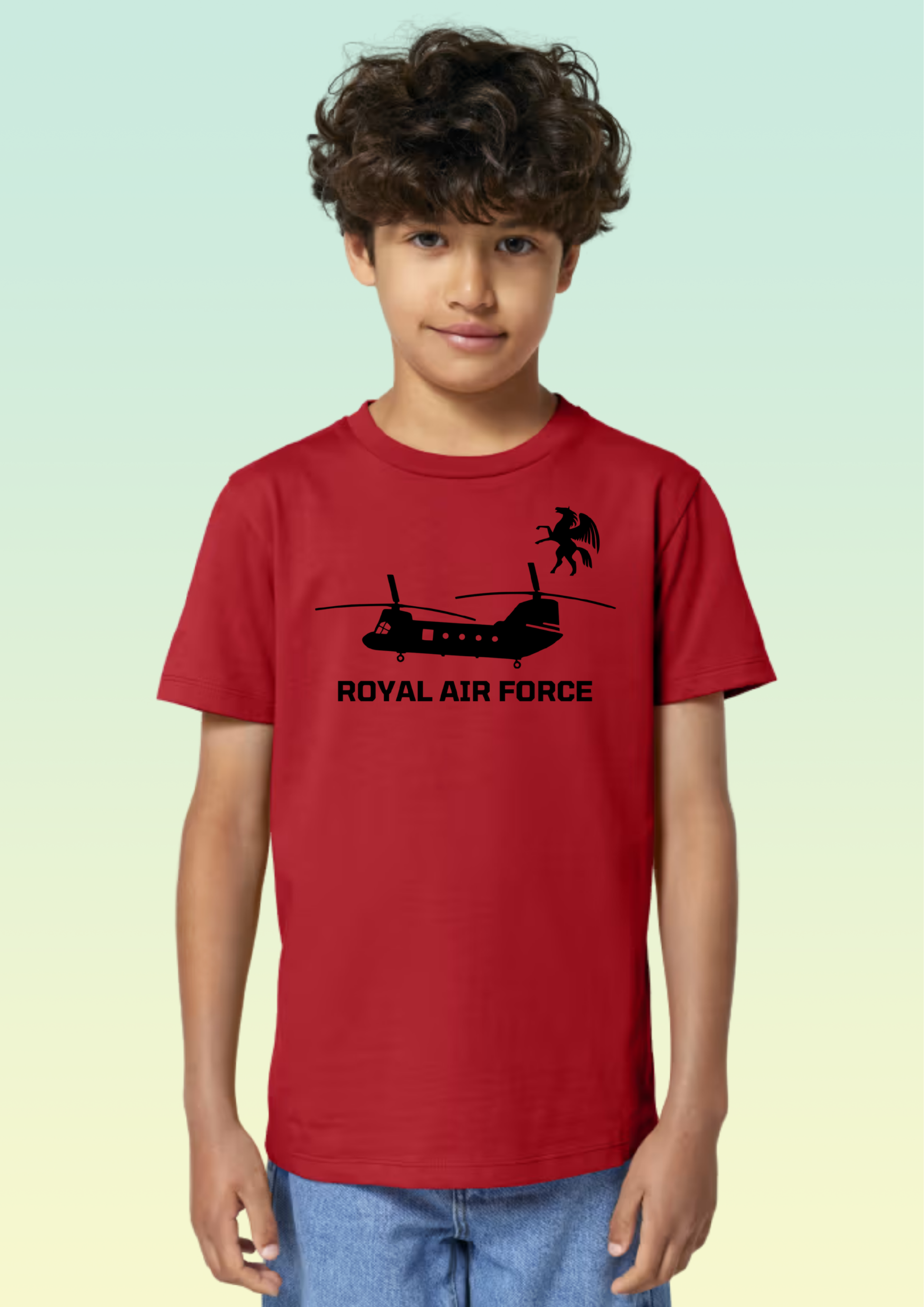 A young boy with curly brown hair wearing a red short-sleeve t-shirt with a black Royal Air Force helicopter graphic and text, paired with blue jeans, set against a light green and yellow gradient background. Exclusive to Flying Teez.
