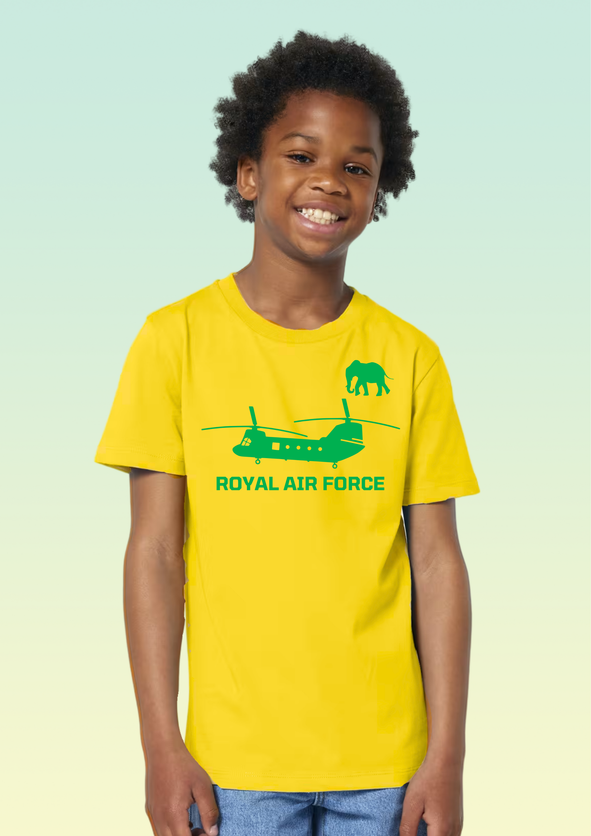 A smiling young boy with curly dark hair stands against a pale mint-to-light-green gradient background wearing a bright yellow short-sleeve T-shirt with a green Royal Air Force helicopter and elephant graphic and the words 'ROYAL AIR FORCE' on the front, paired with blue jeans. Exclusive to Flying Teez.