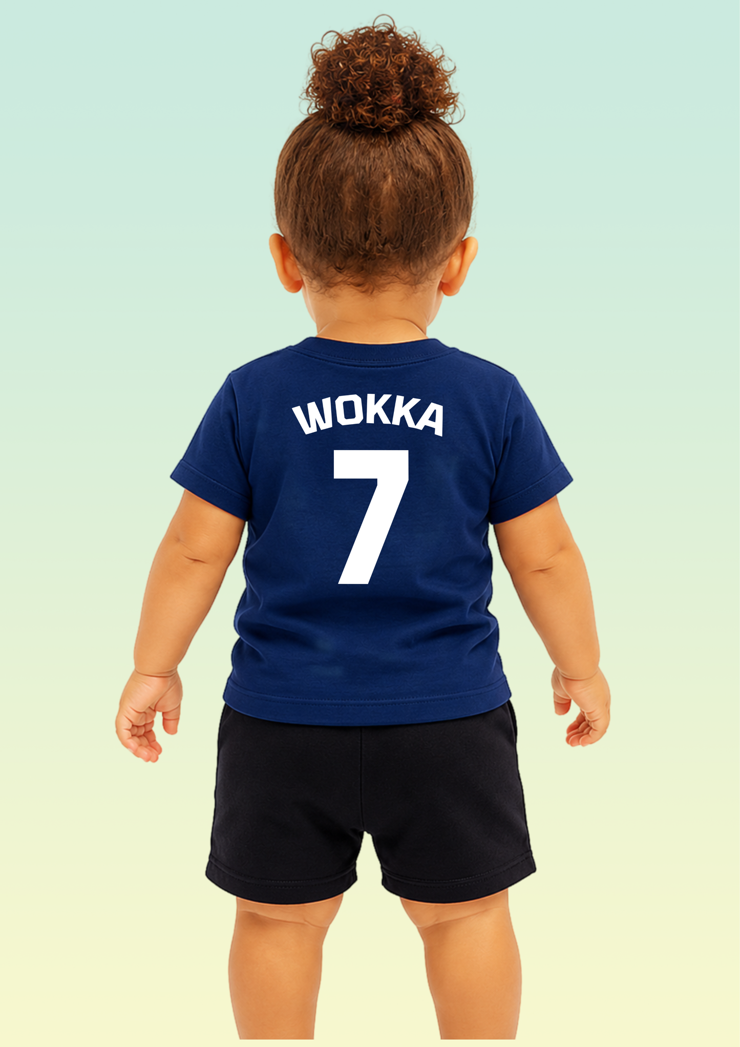 A toddler viewed from behind wearing a navy blue short-sleeve T-shirt with the name 'WOKKA' and number '7' printed in white on the back, paired with dark shorts against a pale gradient background. Exclusive to Flying Teez.