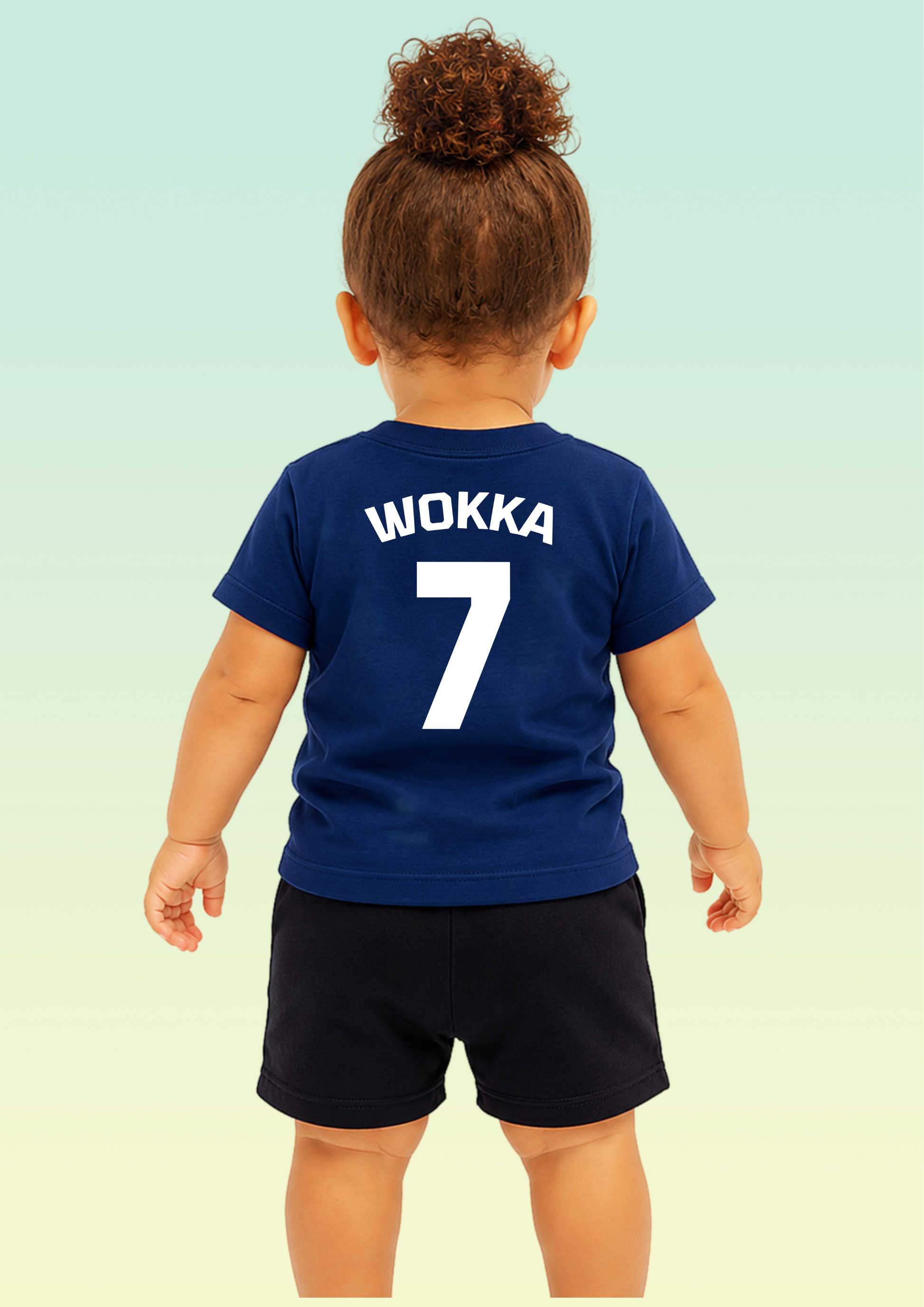 A toddler viewed from behind wearing a navy blue short-sleeve T-shirt with the name 'WOKKA' and number '7' printed in white on the back, paired with dark shorts against a pale gradient background. Exclusive to Flying Teez.