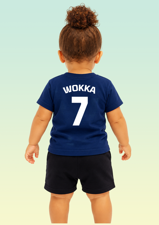 A toddler viewed from behind wearing a navy blue short-sleeve T-shirt with the name 'WOKKA' and number '7' printed in white on the back, paired with dark shorts against a pale gradient background. Exclusive to Flying Teez.