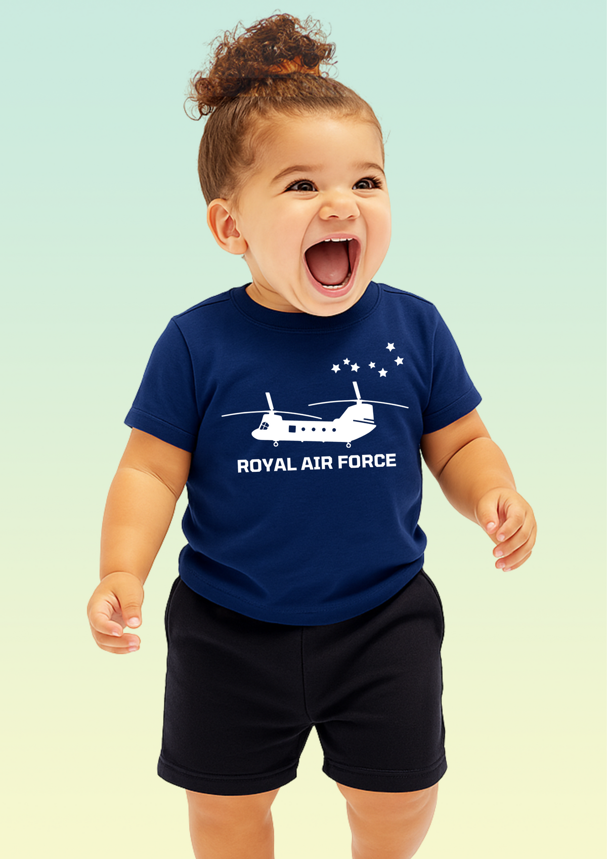 A smiling toddler wearing a navy blue short-sleeve t-shirt with a white Royal Air Force helicopter graphic and text, paired with black shorts, set against a soft light green gradient background. Exclusive to Flying Teez.