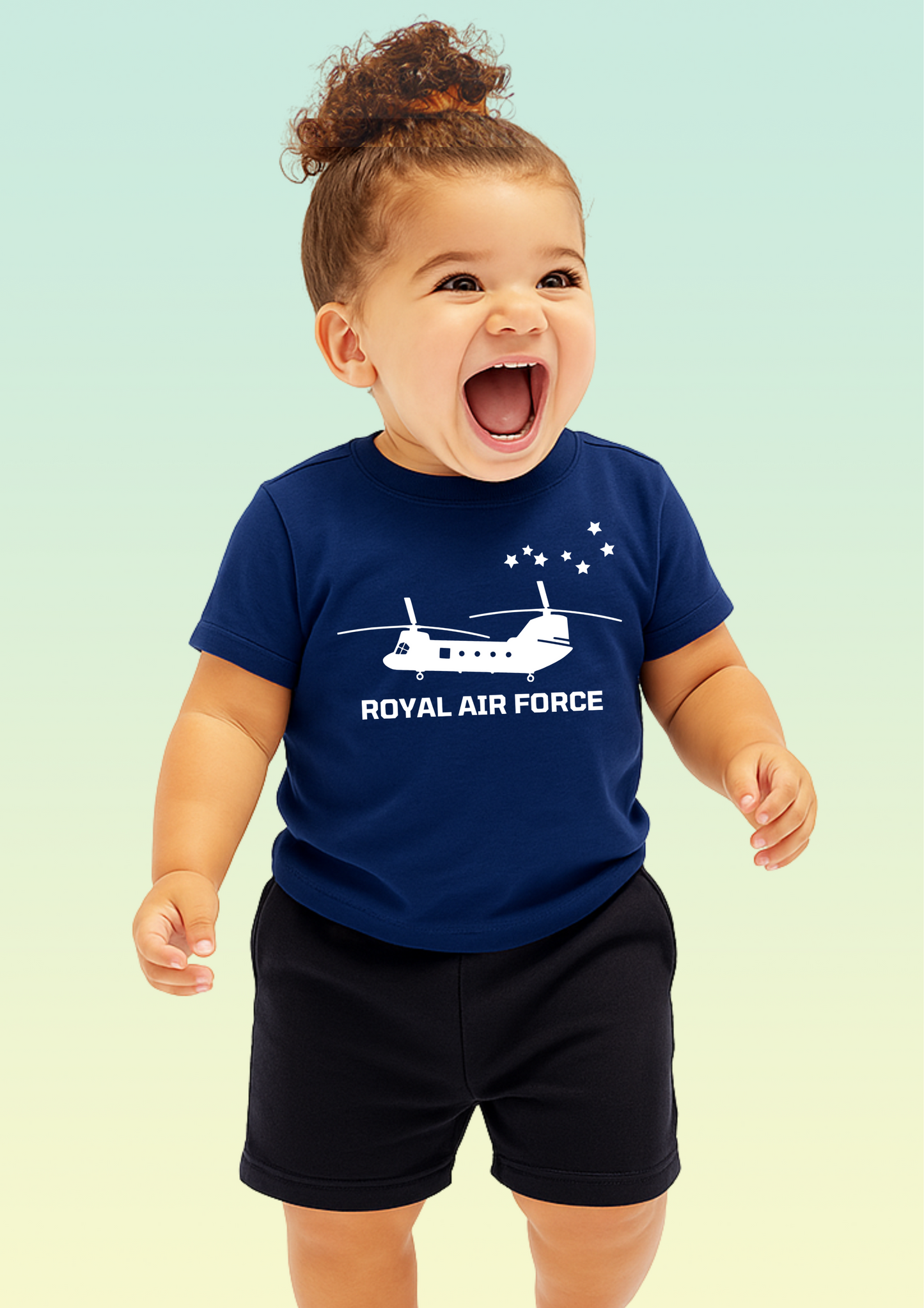 A smiling toddler wearing a navy blue short-sleeve t-shirt with a white Royal Air Force helicopter graphic and text, paired with black shorts, set against a soft light green gradient background. Exclusive to Flying Teez.