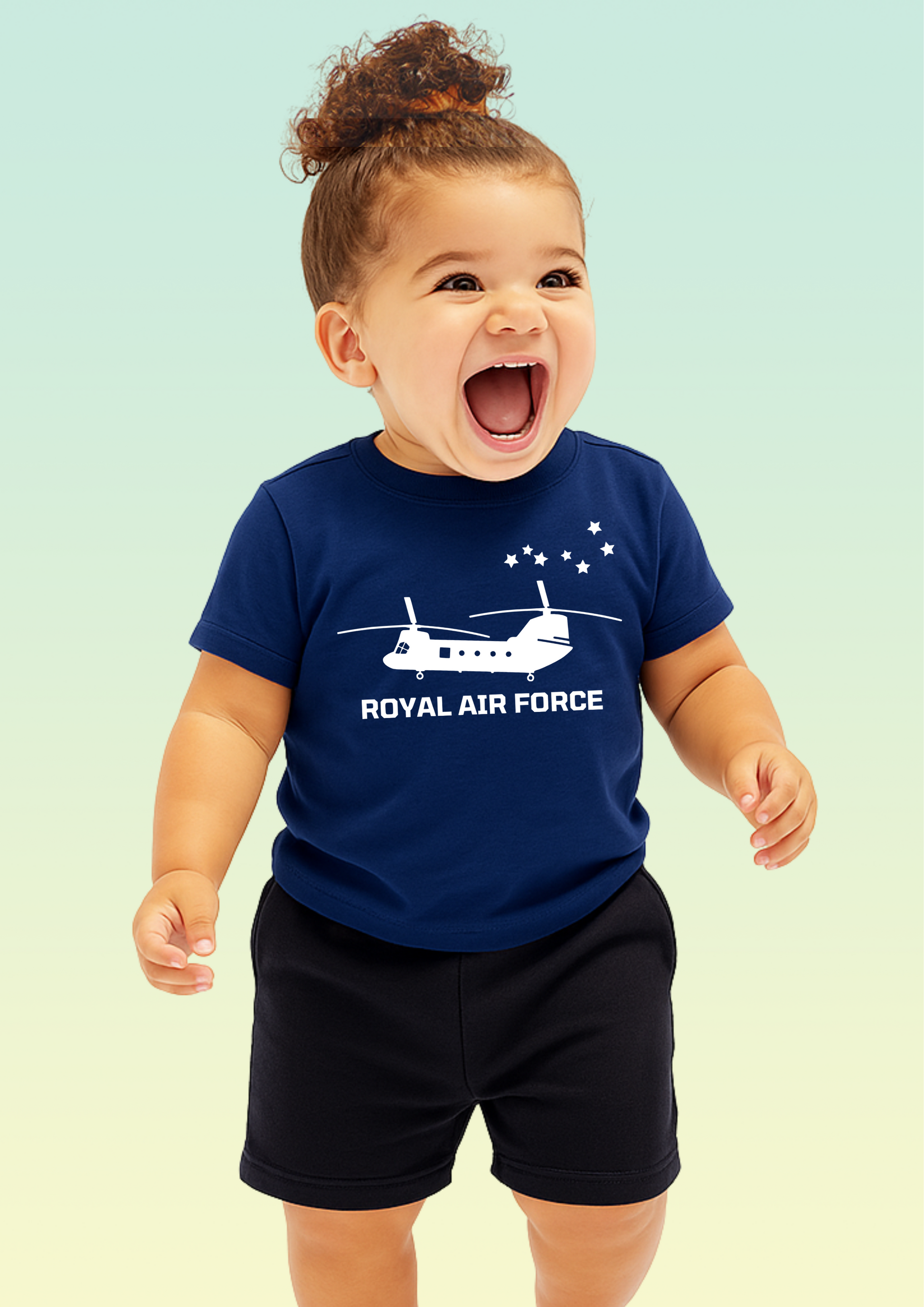 A smiling toddler wearing a navy blue short-sleeve t-shirt with a white Royal Air Force helicopter graphic and text, paired with black shorts, set against a soft light green gradient background. Exclusive to Flying Teez.