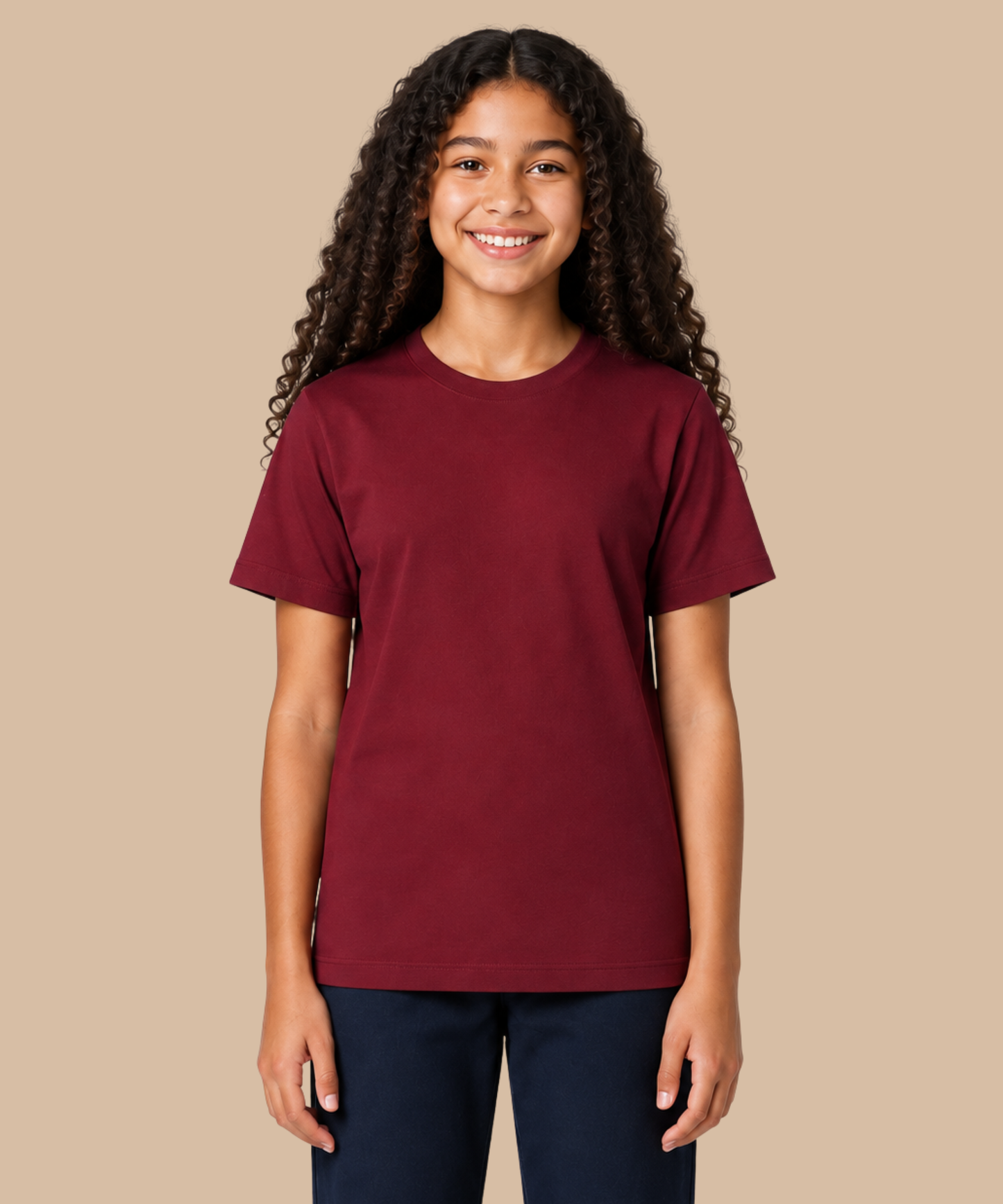 A smiling young woman with long curly dark hair wearing a solid maroon short-sleeve crewneck t-shirt and dark navy pants, standing against a solid beige background. Handprinted t-shirt services from Pathfinder Printworks.