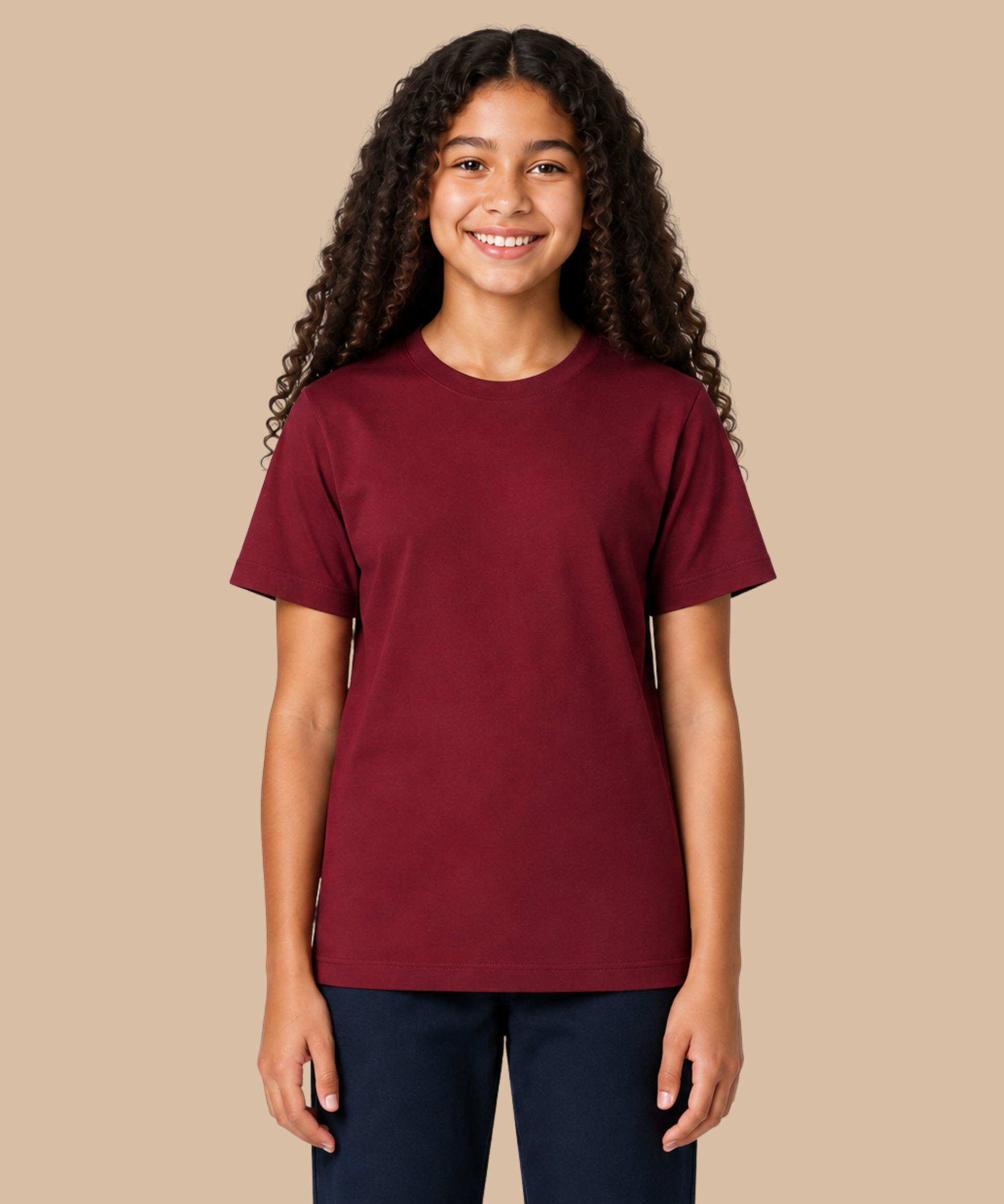A smiling young woman with long curly dark hair wearing a solid maroon short-sleeve crewneck t-shirt and dark navy pants, standing against a solid beige background. Handprinted t-shirt services from Pathfinder Printworks.