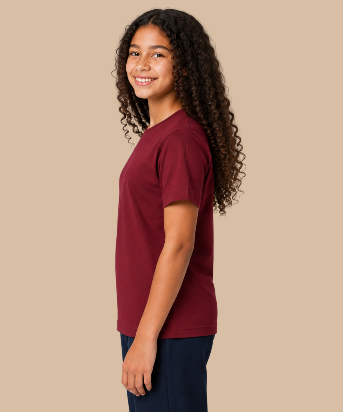A young girl with long, curly dark hair is shown in profile wearing a short-sleeve crewneck t-shirt in maroon and dark pants, standing against a solid beige studio background. Handprinted t-shirt services from Pathfinder Printworks.