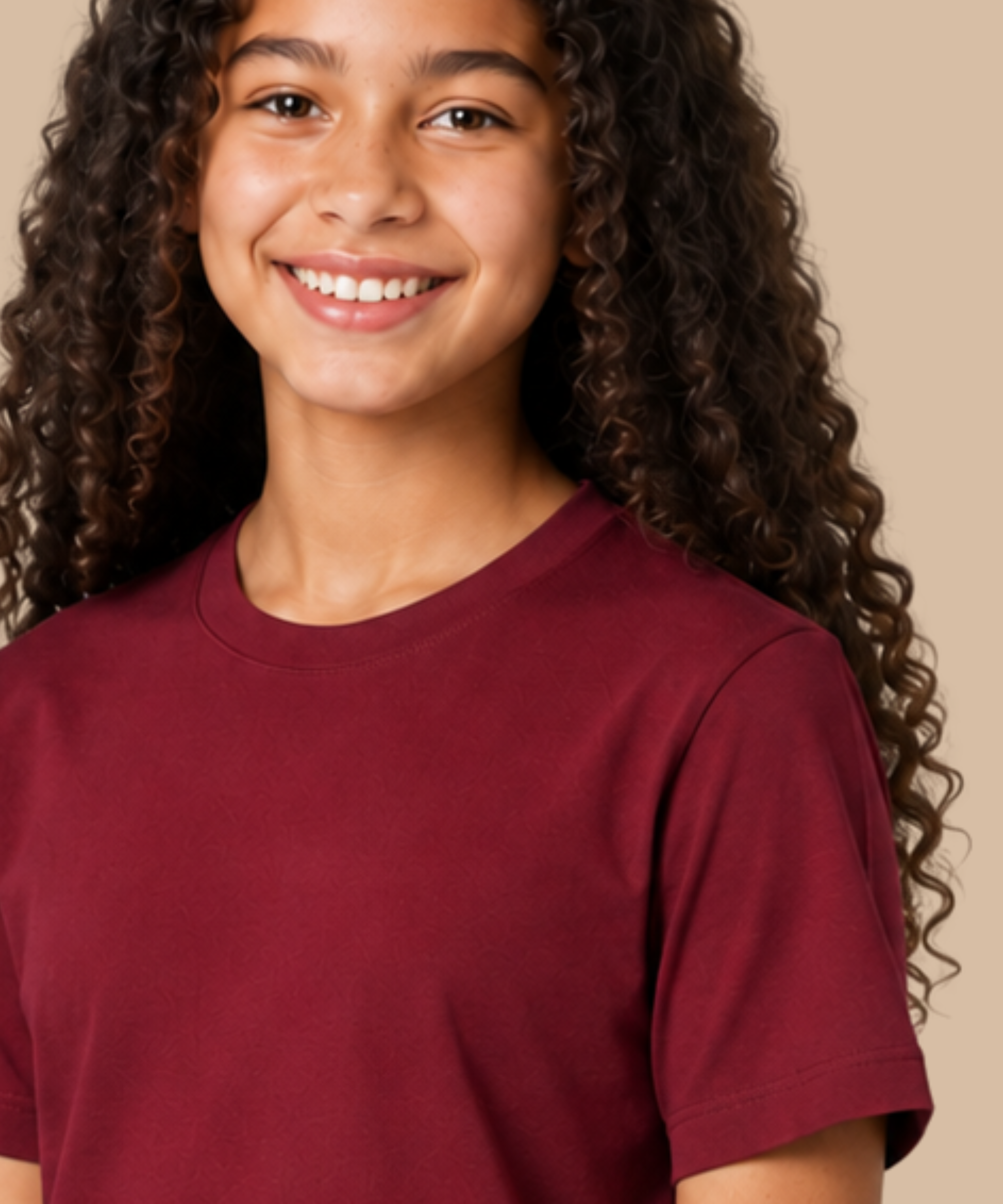A smiling young woman with long, curly dark brown hair wearing a solid maroon crew-neck t-shirt, photographed against a solid beige background. Handprinted t-shirt services from Pathfinder Printworks.