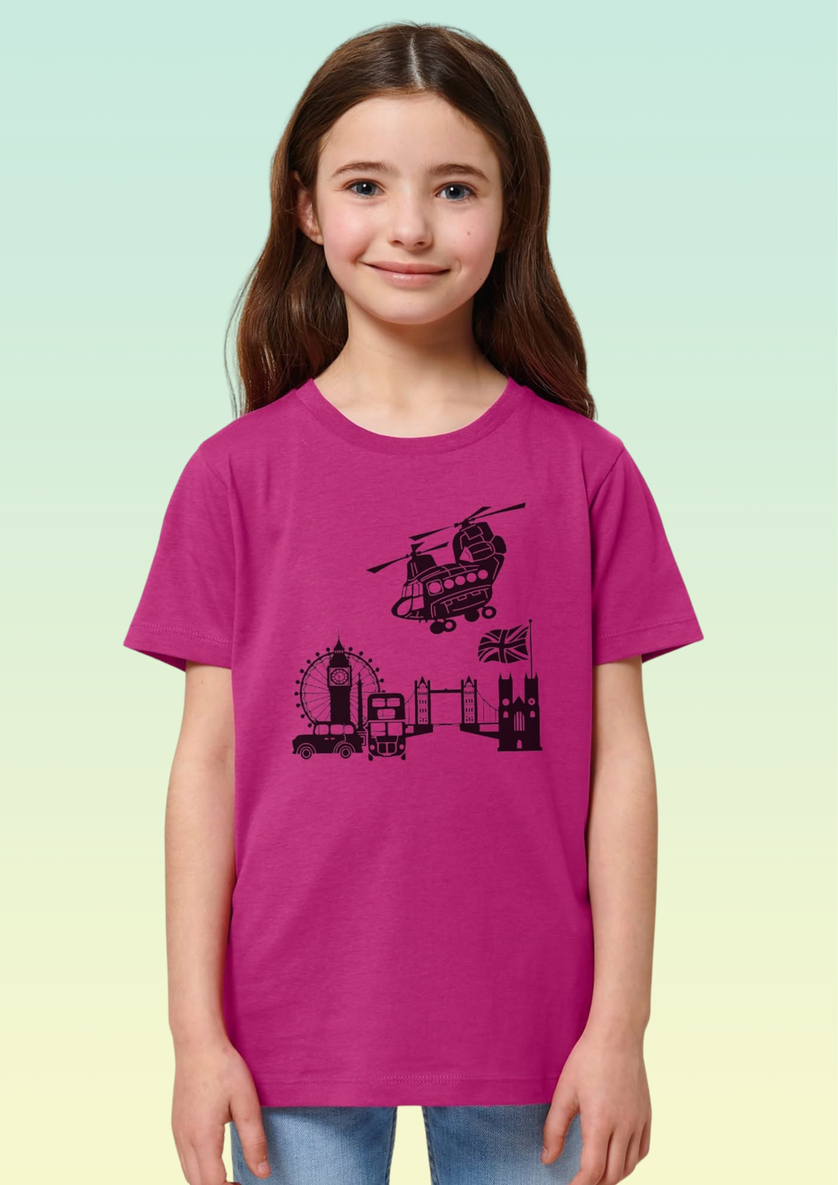 A young girl with long brown hair stands against a light green-to-yellow gradient background wearing a bright magenta t-shirt with a black graphic of London landmarks, including Big Ben, the London Eye, and a helicopter. Exclusive to Flying Teez.