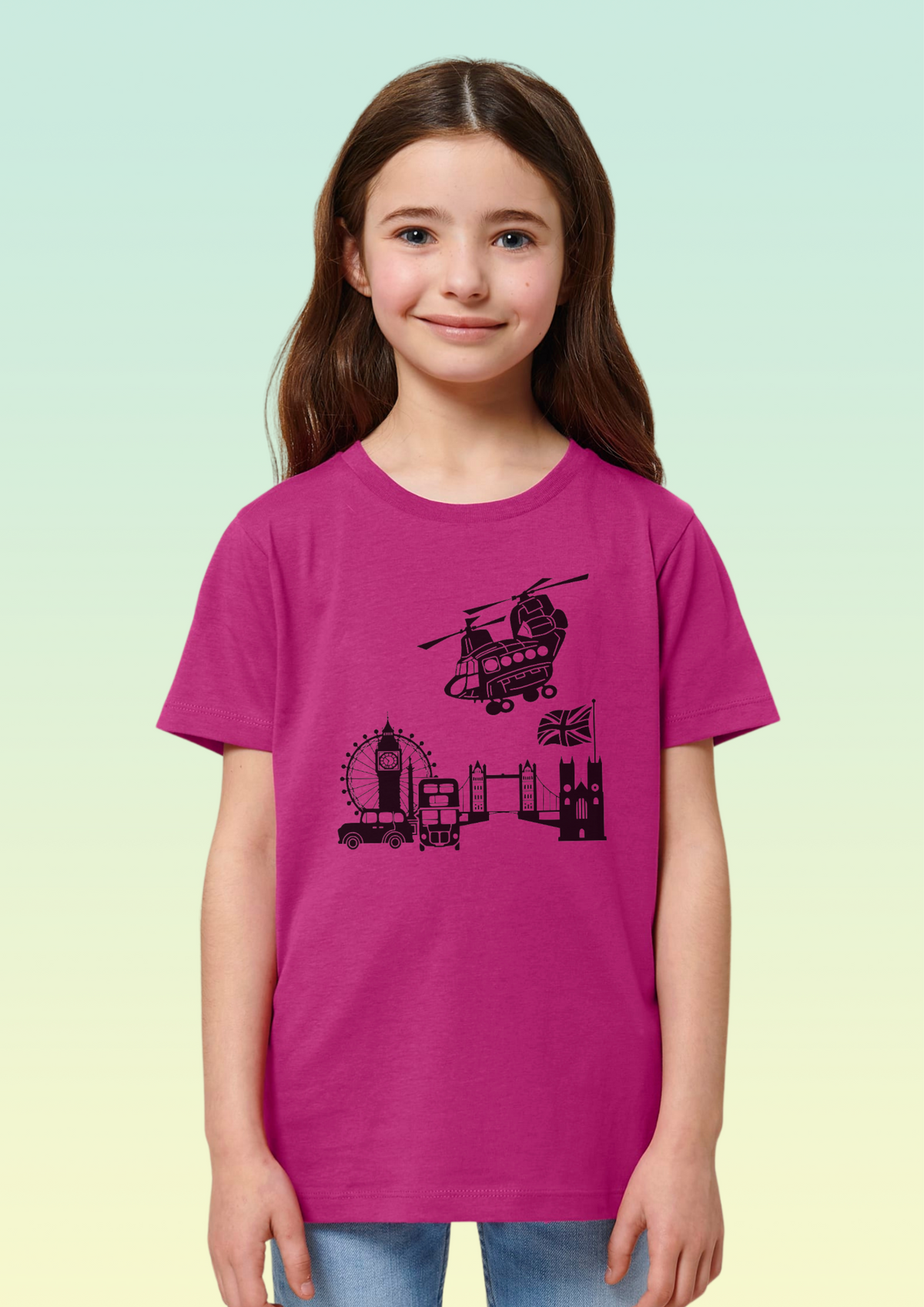 A young girl with long brown hair stands against a light green-to-yellow gradient background wearing a bright magenta t-shirt with a black graphic of London landmarks, including Big Ben, the London Eye, and a helicopter. Exclusive to Flying Teez.