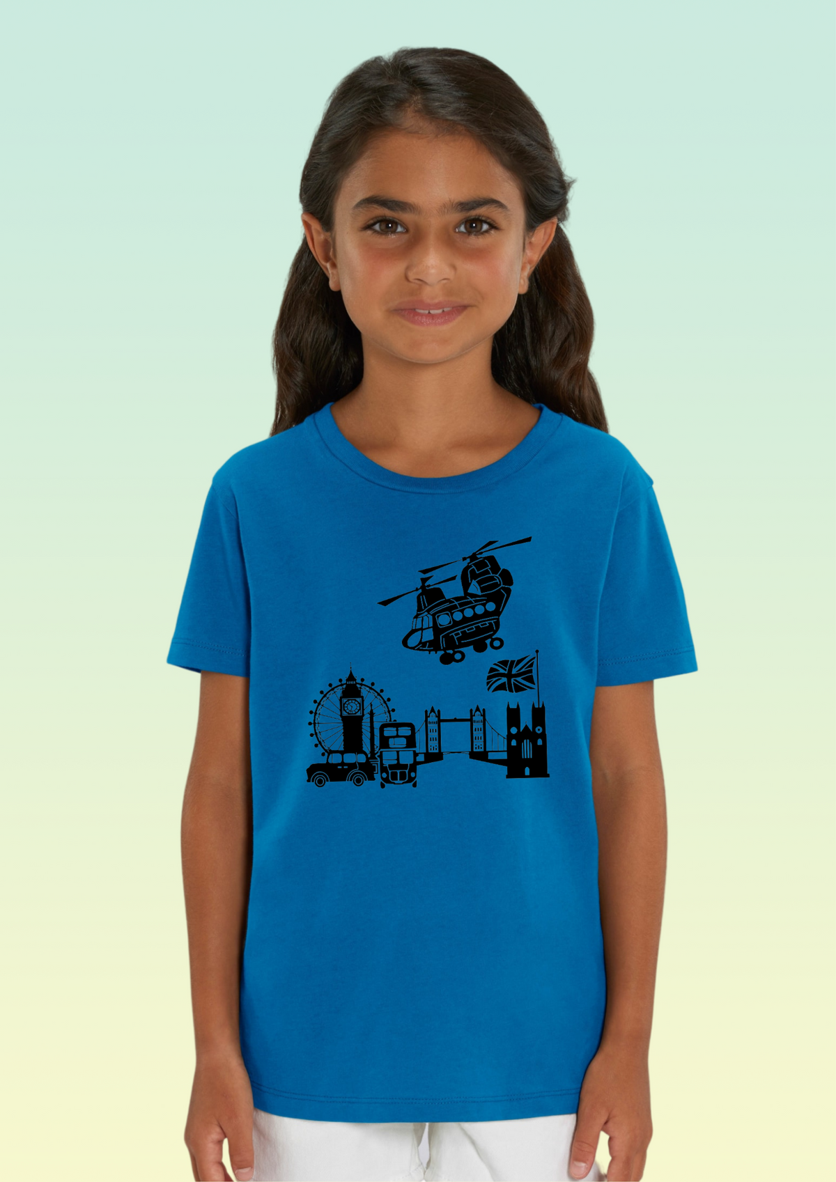 A young girl with dark hair wearing a bright blue short-sleeve t-shirt with a black graphic print of London landmarks, set against a light green and yellow gradient background. Exclusive to Flying Teez.