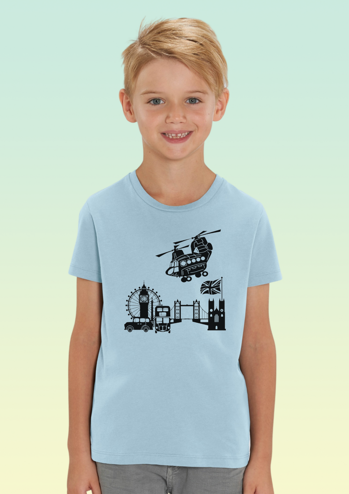 A young boy with blonde hair smiling, wearing a light blue crewneck t-shirt with a black graphic of London landmarks (Big Ben, London Eye, Tower Bridge, Westminster Bridge, and a helicopter). The background is a soft green-to-yellow gradient. Exclusive to Flying Teez.