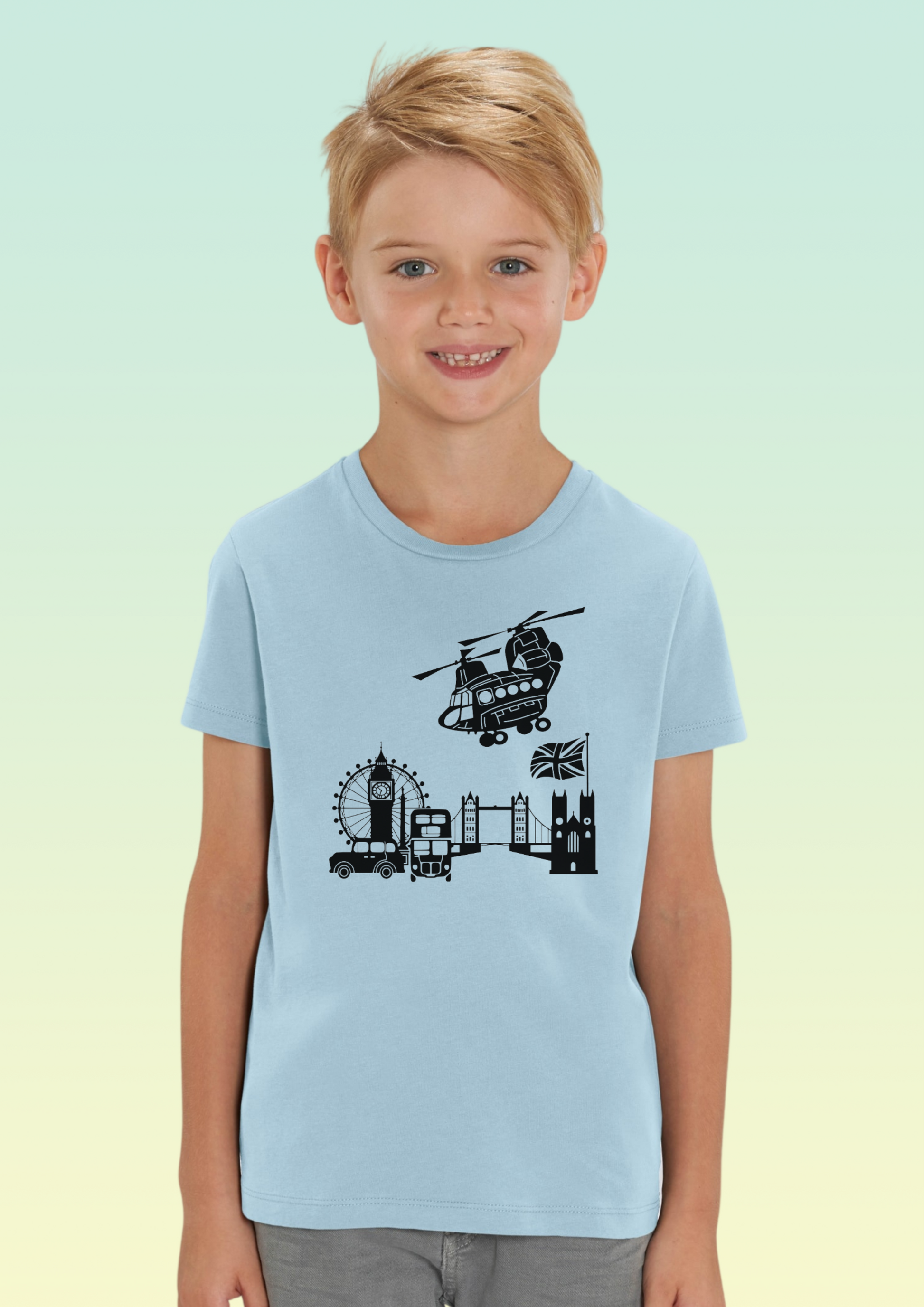 A young boy with blonde hair smiling, wearing a light blue crewneck t-shirt with a black graphic of London landmarks (Big Ben, London Eye, Tower Bridge, Westminster Bridge, and a helicopter). The background is a soft green-to-yellow gradient. Exclusive to Flying Teez.