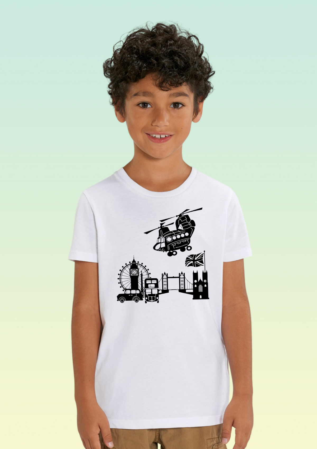 A young boy with curly dark hair wearing a white short-sleeve t-shirt with a black graphic print of London landmarks (Big Ben, London Eye, Tower Bridge, Westminster Abbey, and a helicopter) on the front, set against a light green to pale yellow gradient background. Exclusive to Flying Teez.