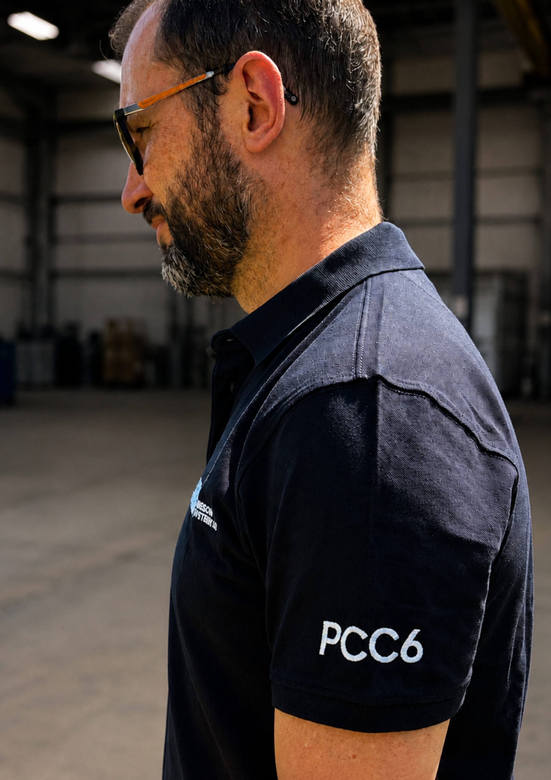 A man in profile wearing a navy short-sleeve polo shirt with 'PCC6' embroidered in white on the sleeve, standing in a warehouse-like environment. Embroidery services from Pathfinder Printworks.