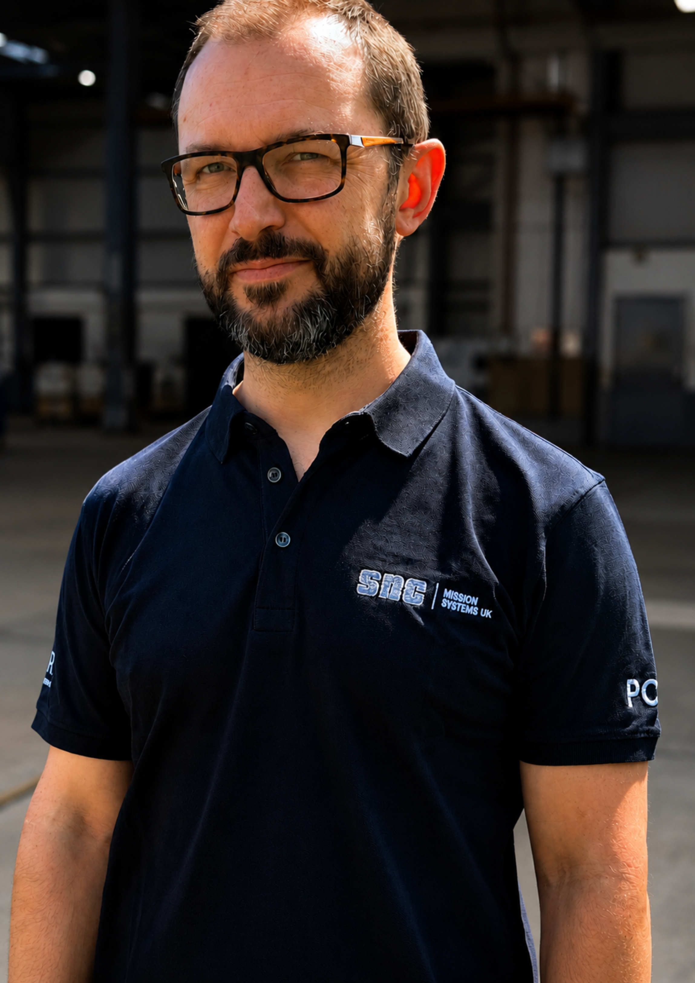 A man with glasses and a beard wearing a dark navy blue polo shirt with 'SNC Mission Systems UK' embroidered on the chest, standing in an industrial setting. Embroidery services from Pathfinder Printworks.