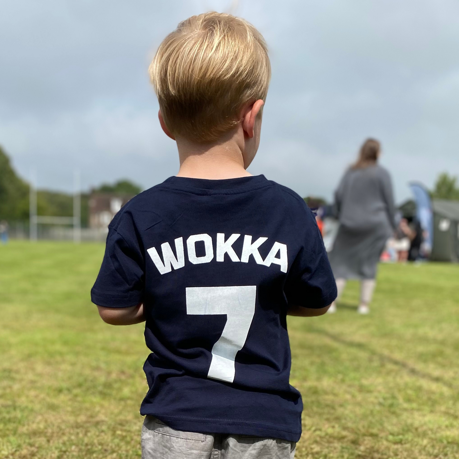 A young boy seen from behind wearing a navy blue t-shirt with 'WOKKA 7' printed in white on the back, standing on a grassy field. Hand printed by Pathfinder Printworks - based in South Wales.