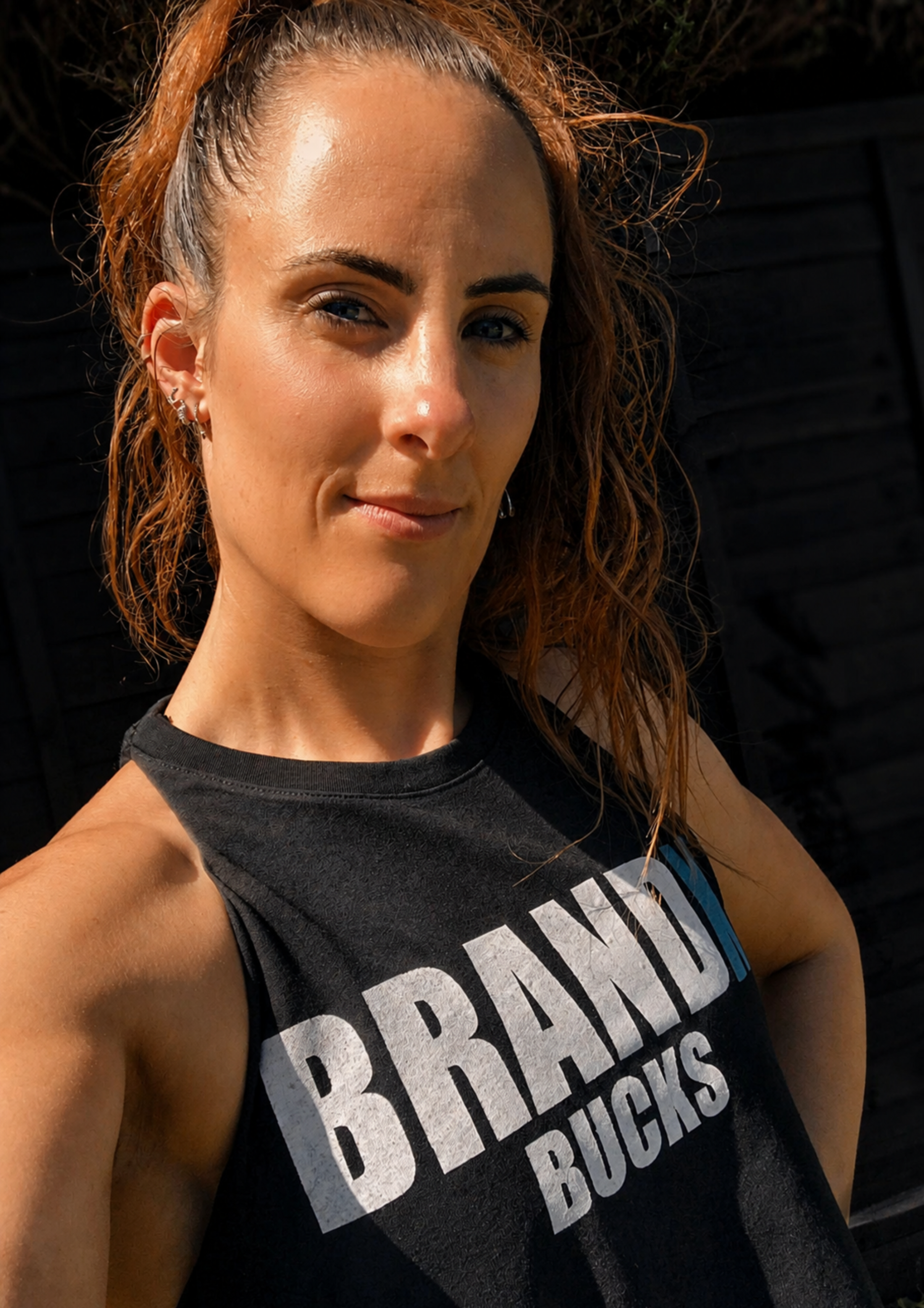 Portrait of a woman outdoors in warm evening sunlight wearing a black sleeveless "Brand Bucks" fitness top. She faces the camera confidently with hands on hips, showcasing the bold printed design.