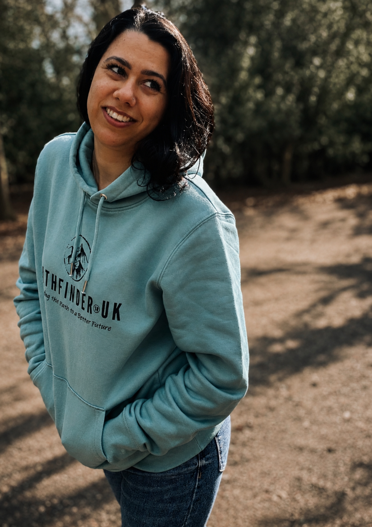 Person wearing a Aloe green Pathfinder UK hoodie standing on a woodland path, smiling while looking off-camera, surrounded by trees and soft natural light.
