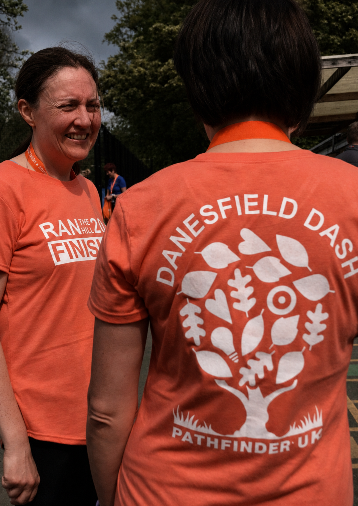 Two participants wearing bright orange event t-shirts chat after completing the Danesfield Dash. One shirt shows the front event print while the other displays a large tree-themed event design on the back.