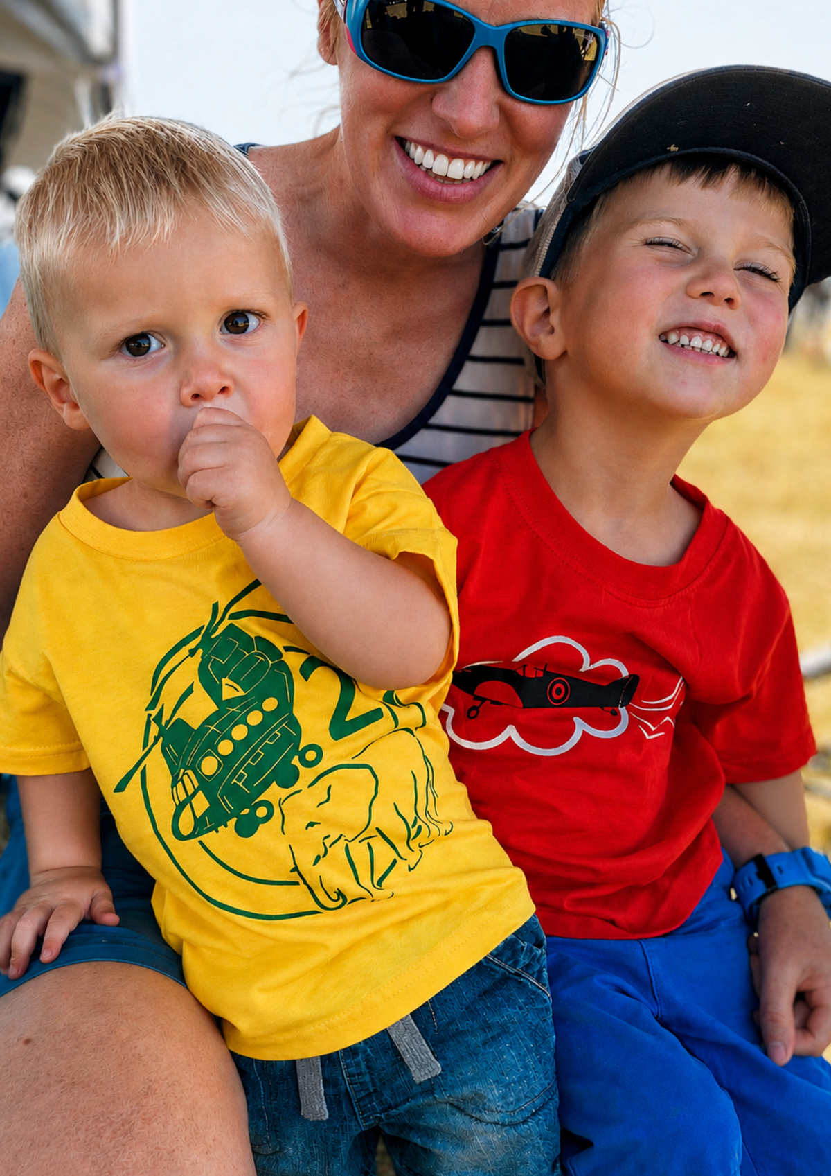 Two young boys wearing colorful graphic t-shirts, with a woman smiling in the background. The boy on the left wears a bright yellow t-shirt with a green helicopter and elephant graphic, while the boy on the right wears a bright red t-shirt with a black and white cloud graphic.