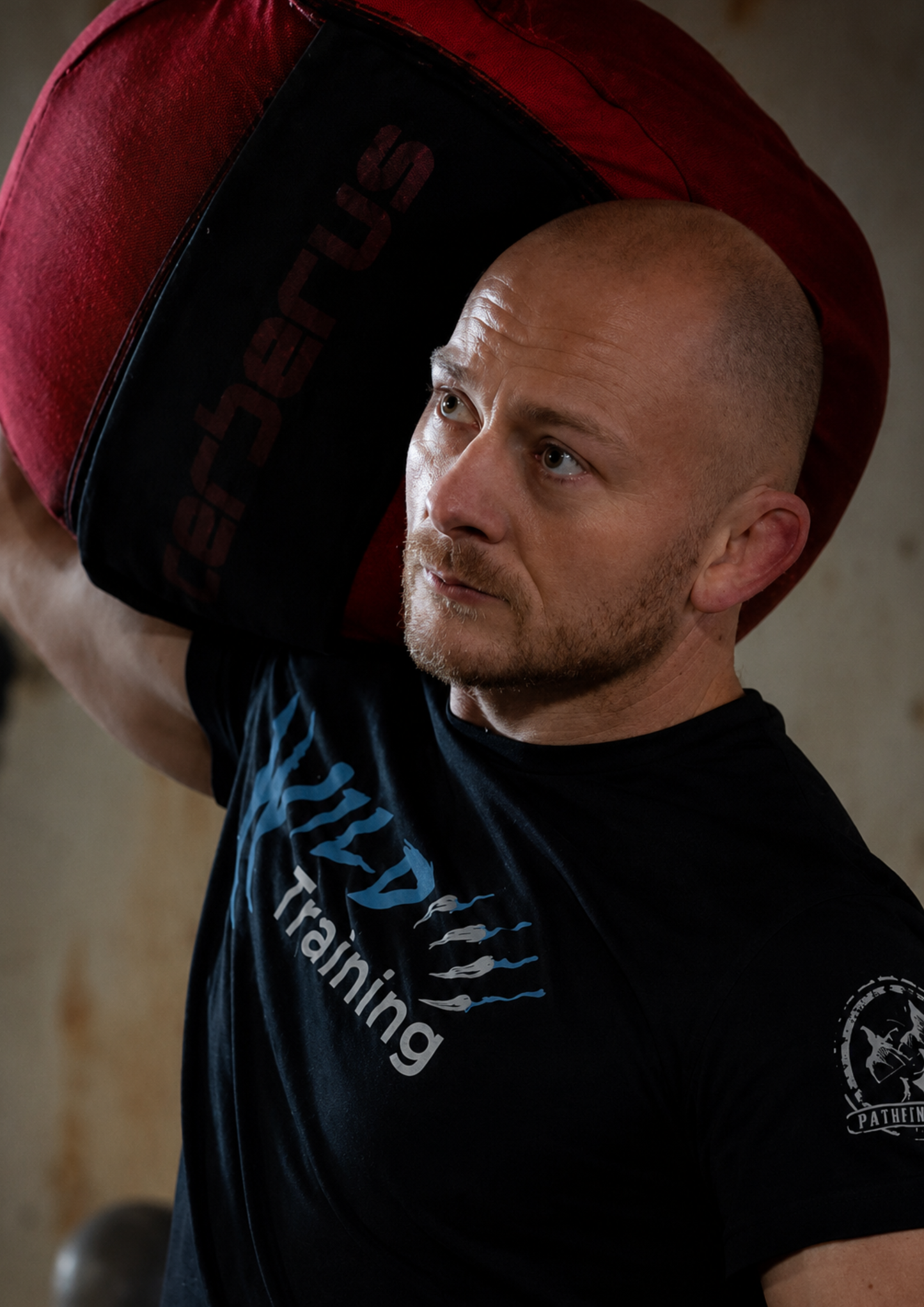 Portrait of a man carrying a heavy sandbag on his shoulder during functional fitness training. The dramatic lighting and focused expression capture the determination, resilience and hard work behind athletic performance.