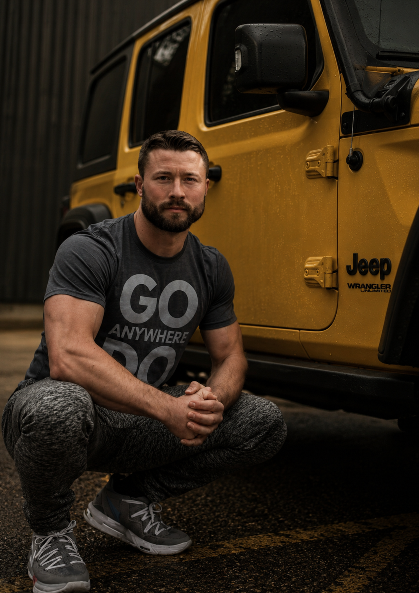 Portrait of a man crouching beside a yellow off-road vehicle, wearing a grey adventure-themed t-shirt. The moody lighting and rugged setting create a strong outdoor lifestyle image focused on exploration and adventure.