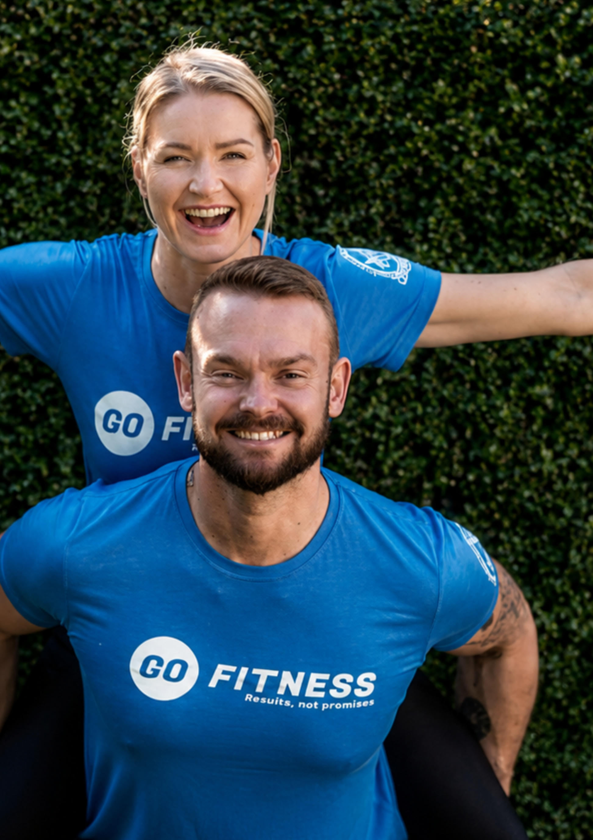 Two fitness coaches wearing matching branded t-shirts smile at the camera, posing playfully together in front of a green backdrop. The image captures personality, teamwork and the approachable side of a fitness business.
