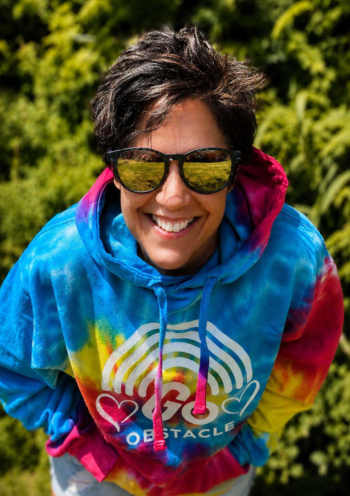 A smiling participant wearing sunglasses and a colourful tie-dye obstacle race hoodie leans towards the camera outdoors. The bright colours and cheerful expression capture the fun, inclusive spirit of community challenge events.