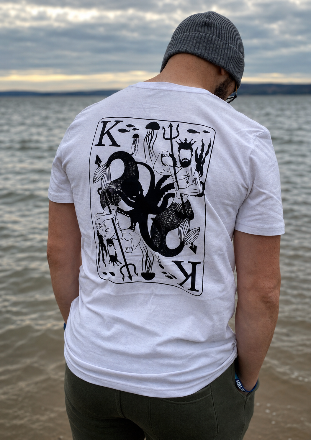 Person standing at the edge of a lake wearing a white t-shirt featuring a large black illustrated King of the Seas playing-card style design on the back. The water and cloudy sky create a dramatic coastal backdrop.