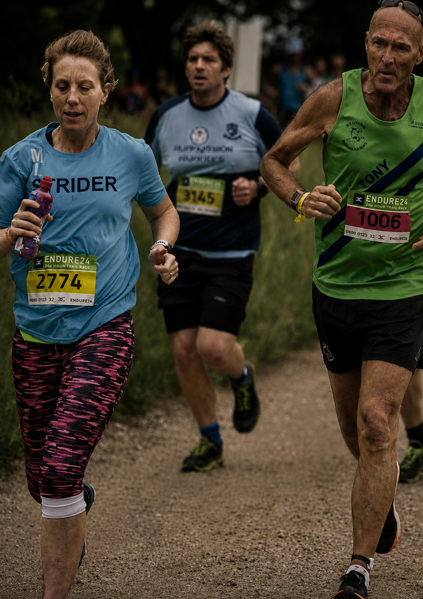 Runners taking part in an endurance trail race on a gravel path. A female runner wearing a light blue event t-shirt is captured in the foreground, with other competitors of different ages running alongside and behind her, illustrating the inclusive nature of endurance events.