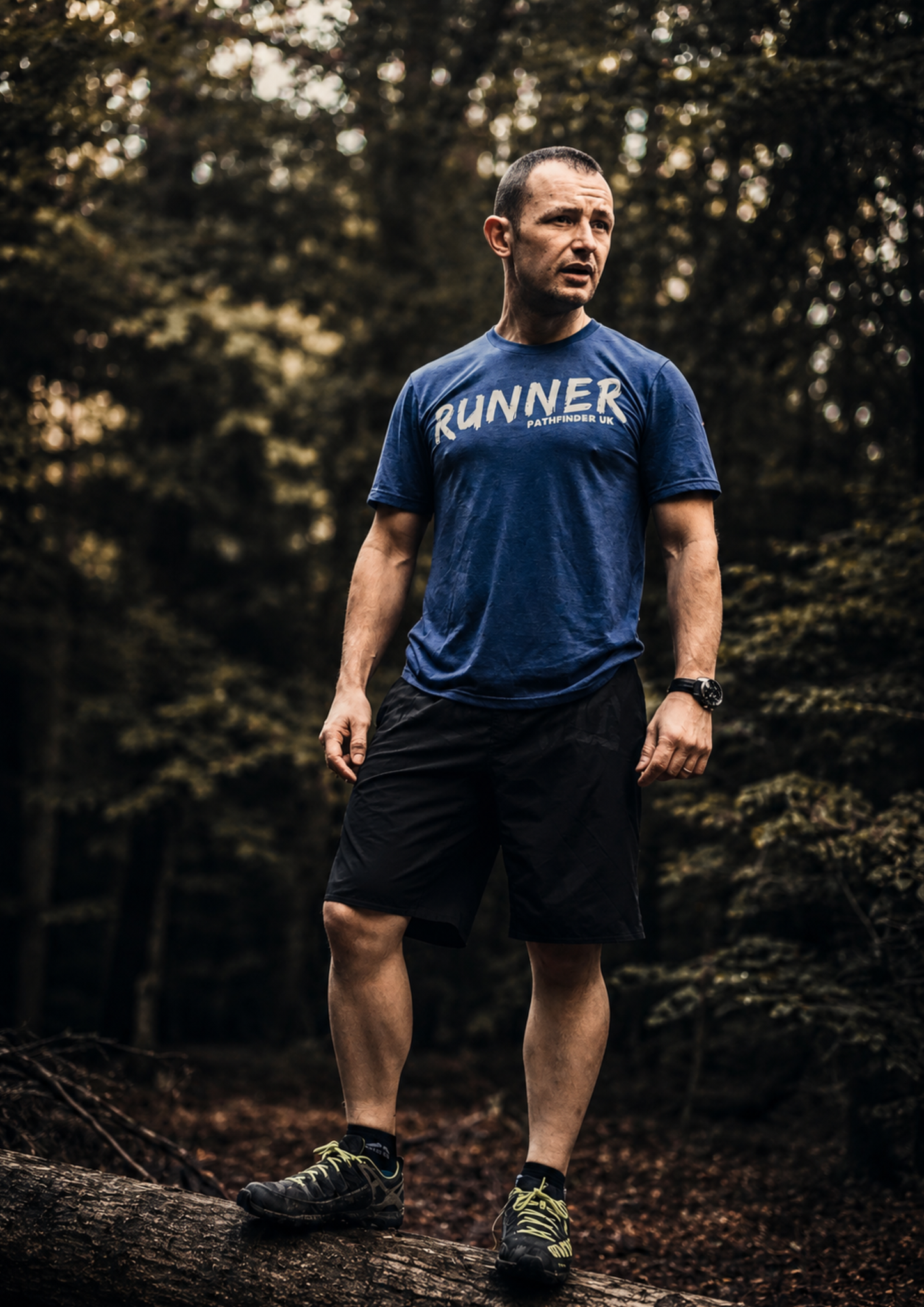 Trail runner standing on a fallen tree in woodland wearing a blue Pathfinder UK t-shirt. The image captures a sense of adventure and exploration, surrounded by dense forest and natural trails.