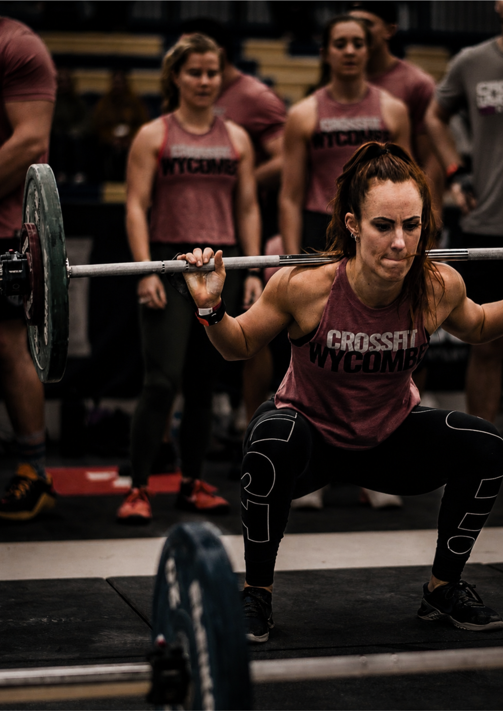 Athlete performing a heavy back squat during a CrossFit competition. Wearing a branded team vest, she is captured at the most demanding point of the lift while teammates watch from the background, highlighting focus, strength and determination.