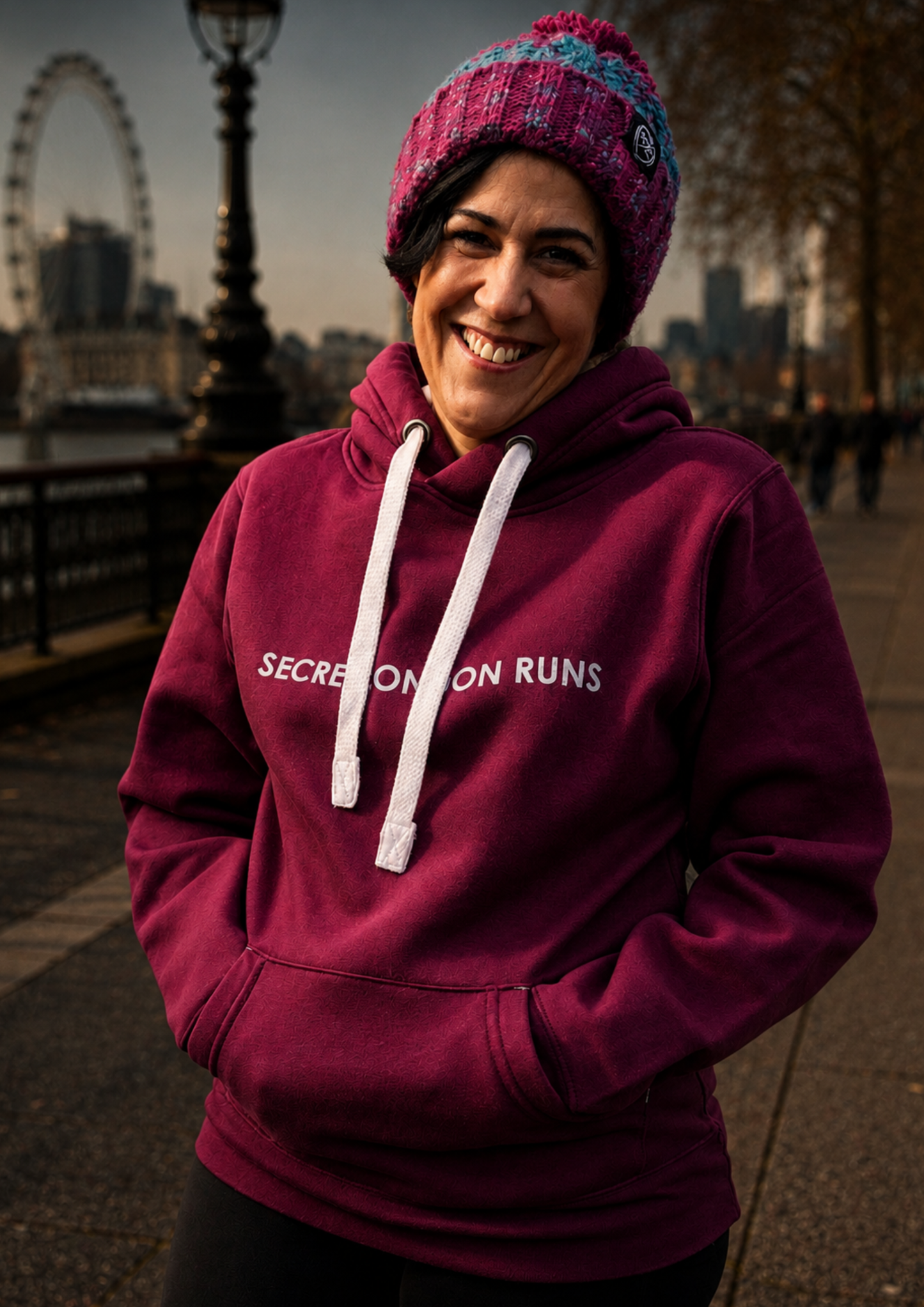 Smiling runner wearing a burgundy Secret London Runs hoodie and knitted hat, standing on a riverside walkway in London with the London Eye visible in the background.