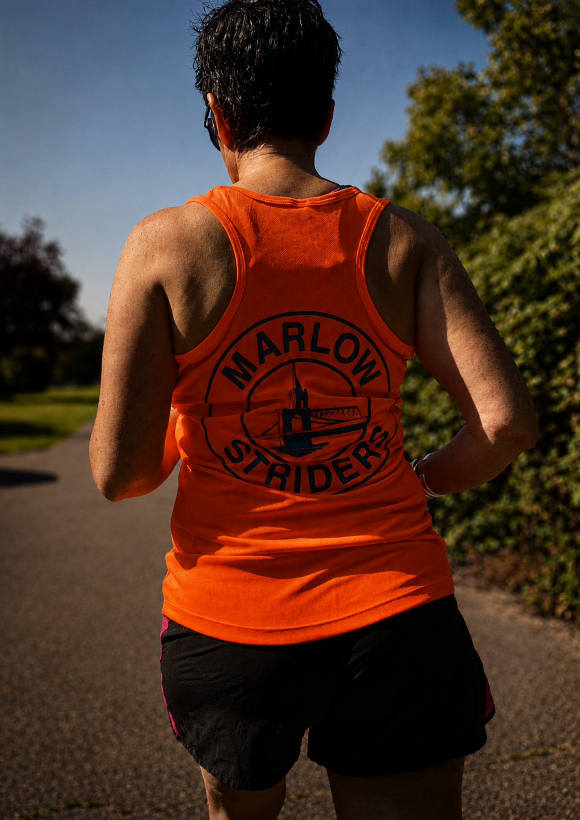 A runner wearing a bright orange running vest with a club logo on the back runs along a quiet country road in warm sunlight. The image captures determination, consistency and the solitary effort behind long-term progress.