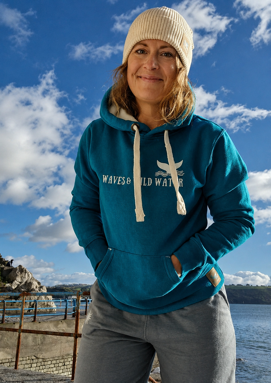 Person standing by the water wearing a turquoise Waves and Wild Water hoodie and cream beanie. The front chest logo is visible against a bright blue sky with scattered clouds and a coastal backdrop.