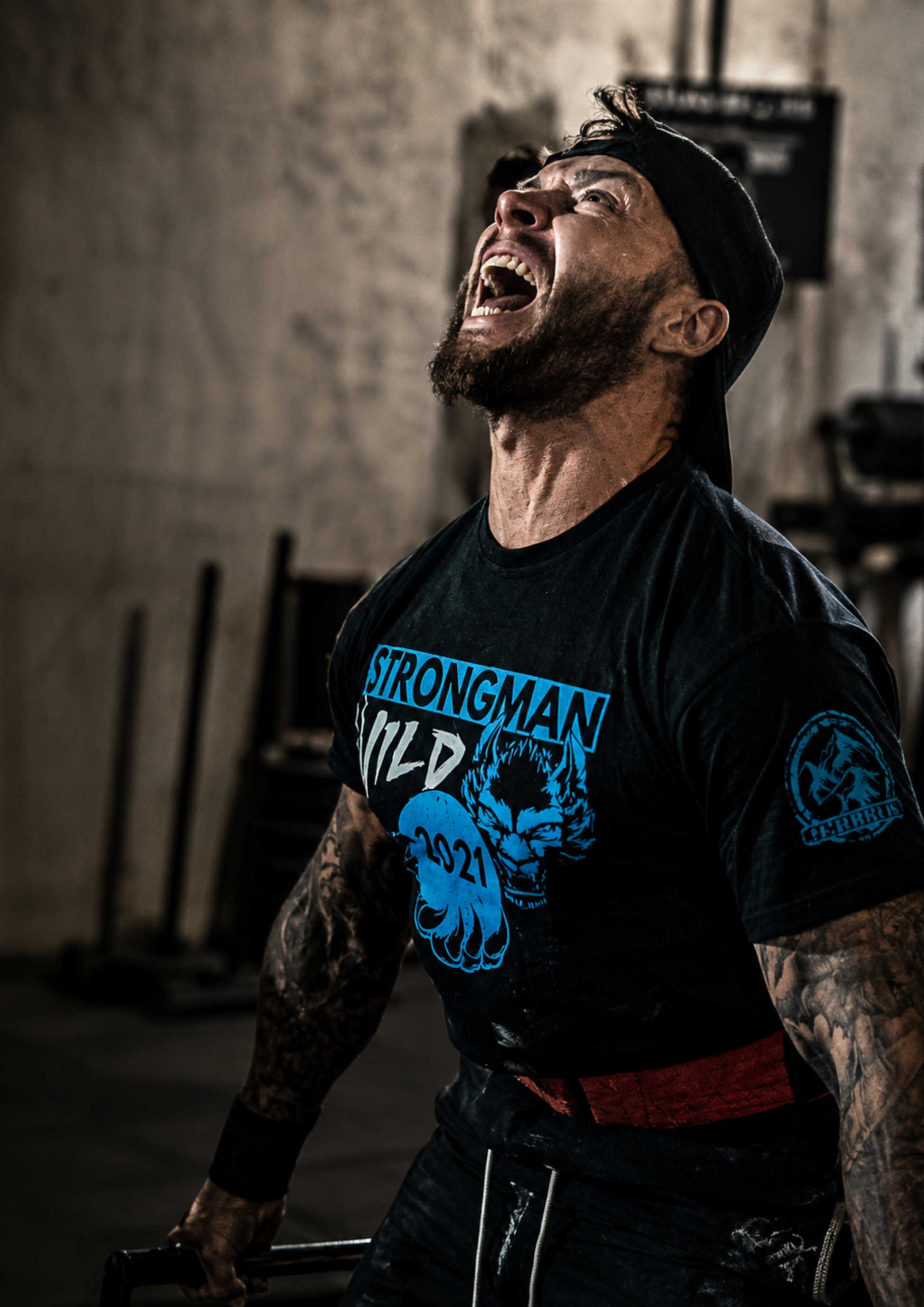 A strongman athlete strains with maximum effort during a lift, his expression showing intensity and determination. Wearing branded competition apparel, he pushes through a challenging moment in a strength training environment.