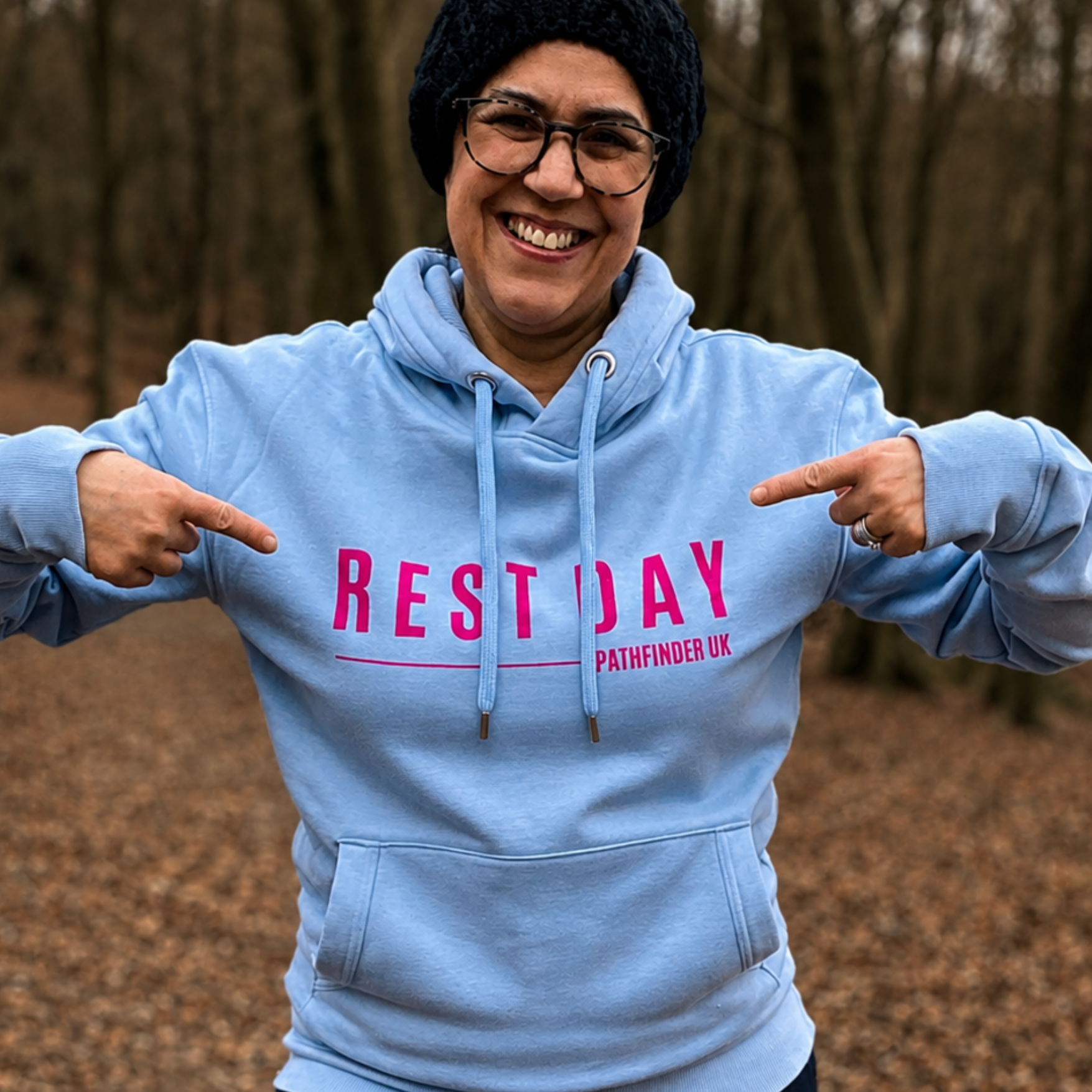 A smiling person wearing a light blue hoodie with pink text reading 'REST DAY' and smaller 'PATHFINDER UK' on the chest, standing outdoors in a wooded setting and pointing at the text.