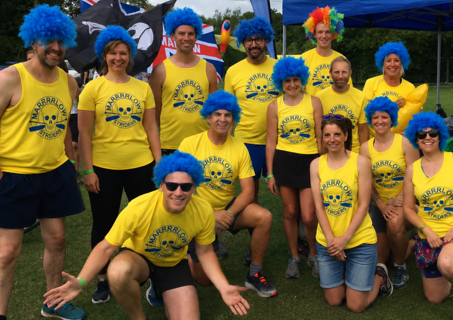 A group wearing matching bright yellow branded shirts and blue wigs pose together at a team event. The image highlights team culture, shared experiences, camaraderie and the fun side of belonging to a community.