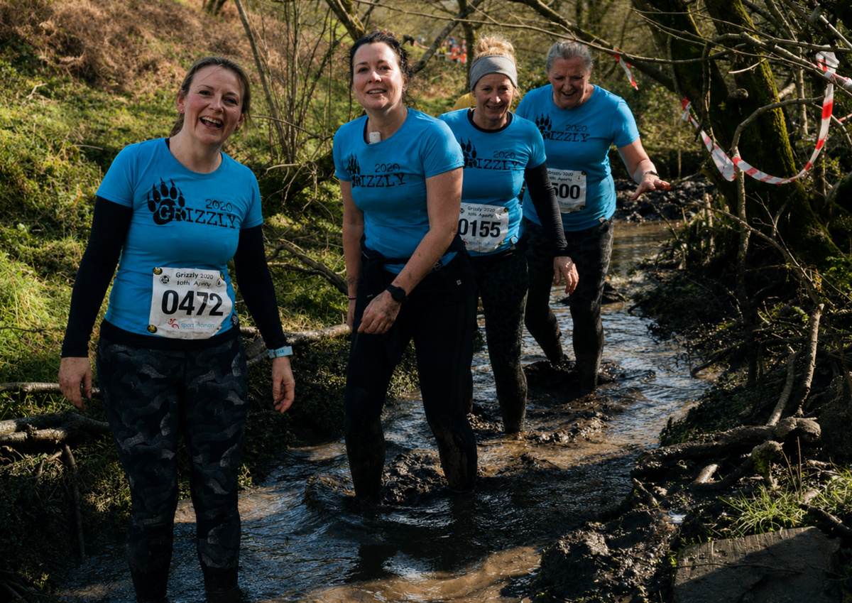 Four runners wearing matching Grizzly event t-shirts walk through a muddy stream during an obstacle race. Despite the challenging conditions, the group are smiling and working their way through the course together, demonstrating teamwork, resilience and camaraderie.