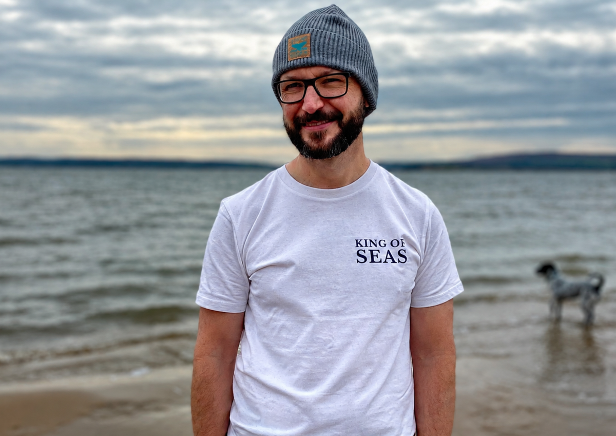 Man wearing a 'King of Seas' t-shirt on a beach with a cloudy sky. Exclusively printed for Wales & Wild Water by Pathfinder Printworks. Hand printed in South Wales.