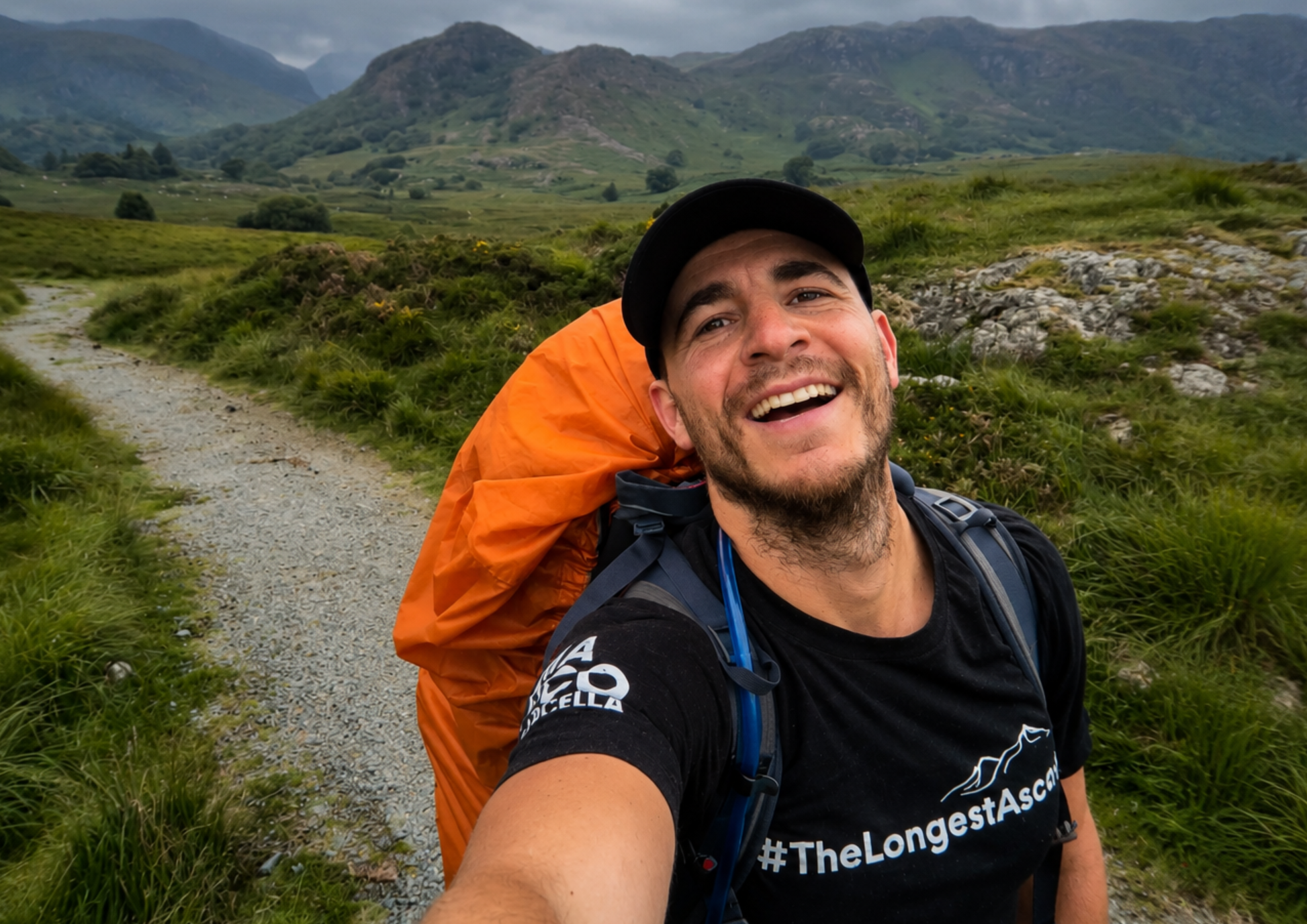 A smiling man taking a selfie outdoors on a trail, wearing a black t-shirt with '#TheLongestAscend' printed on it, a black cap, and a large bright orange hiking backpack. Green hills and mountains are visible in the background under an overcast sky.