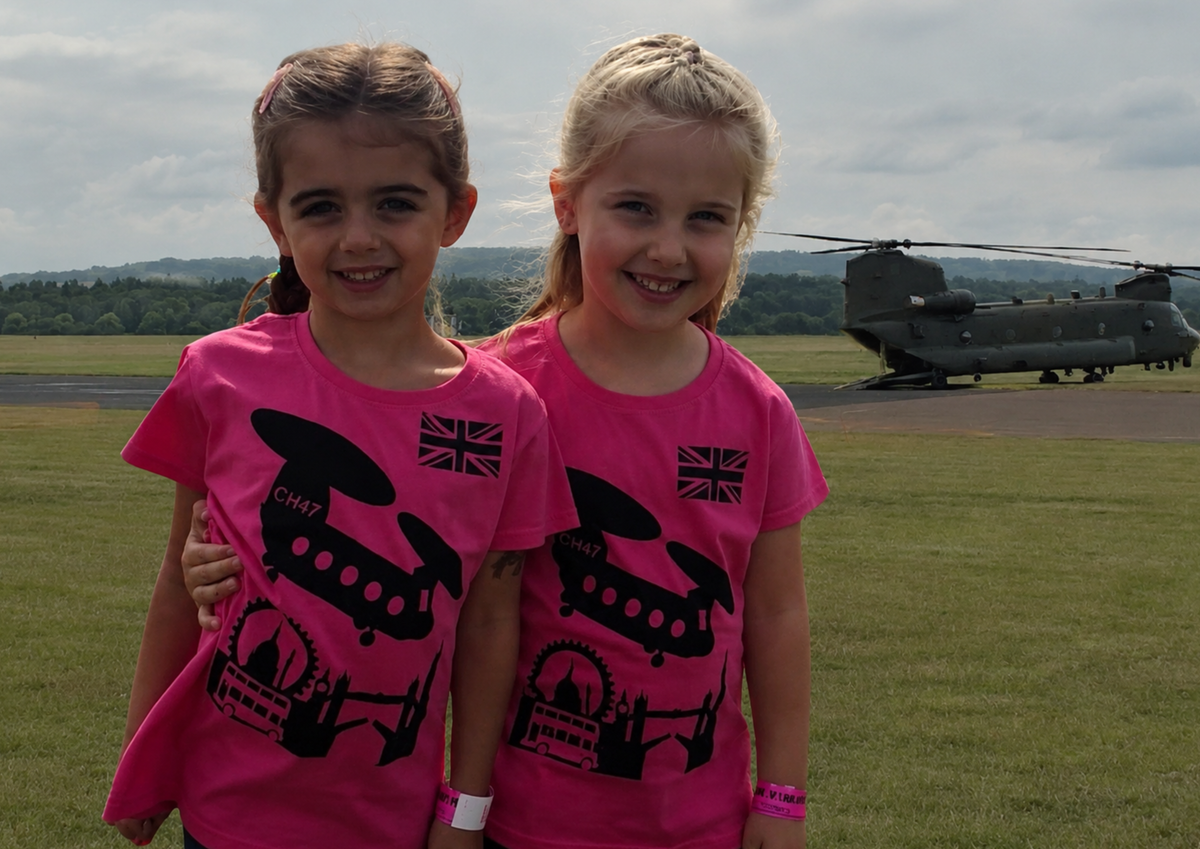 Two girls in pink t-shirts with a helicopter design on a grassy field with a helicopter in the background. Hand printed apparel for teams, clubs and businesses.