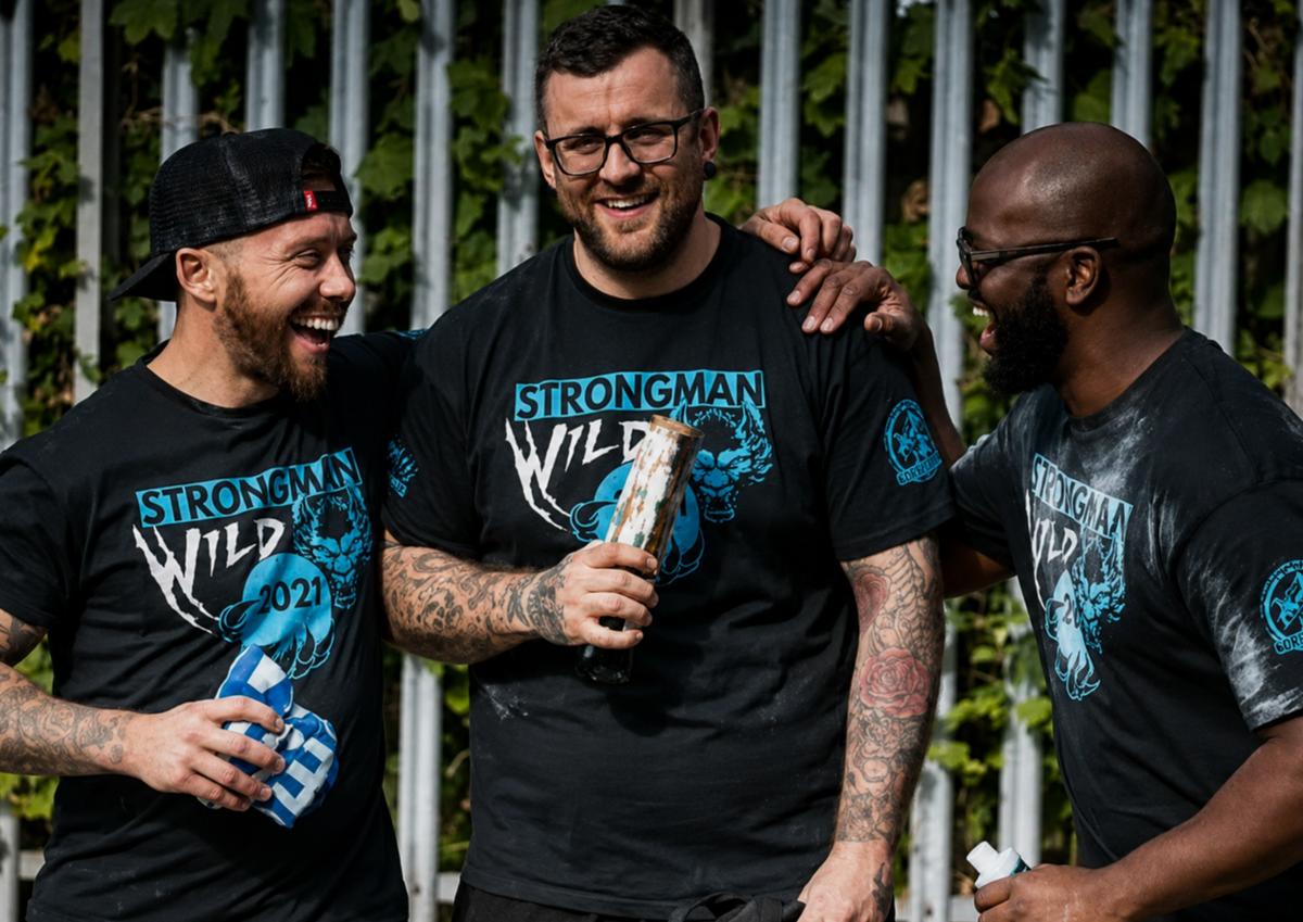 Three men wearing 'Strongman Wild' t-shirts holding bottles, standing in front of a white metal fence. Pathfinder Printworks hand screen print for any organisation. Contact us for details.