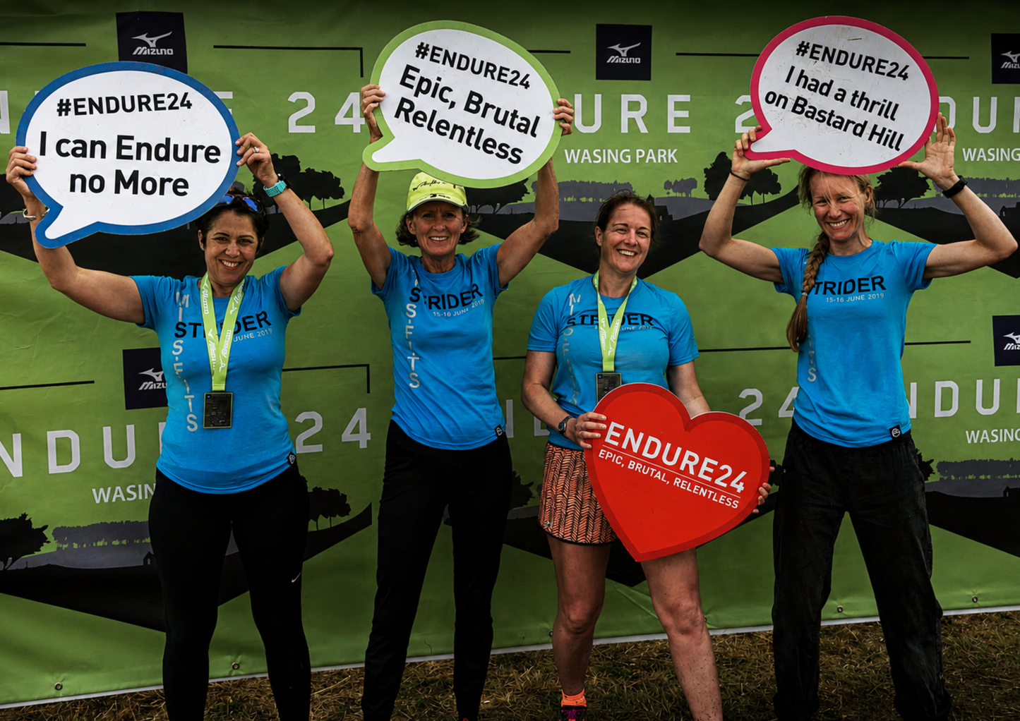 Four women wearing matching blue event t-shirts and medals pose in front of an endurance event backdrop. They hold humorous speech-bubble signs celebrating the challenge while smiling and laughing together after completing the event.