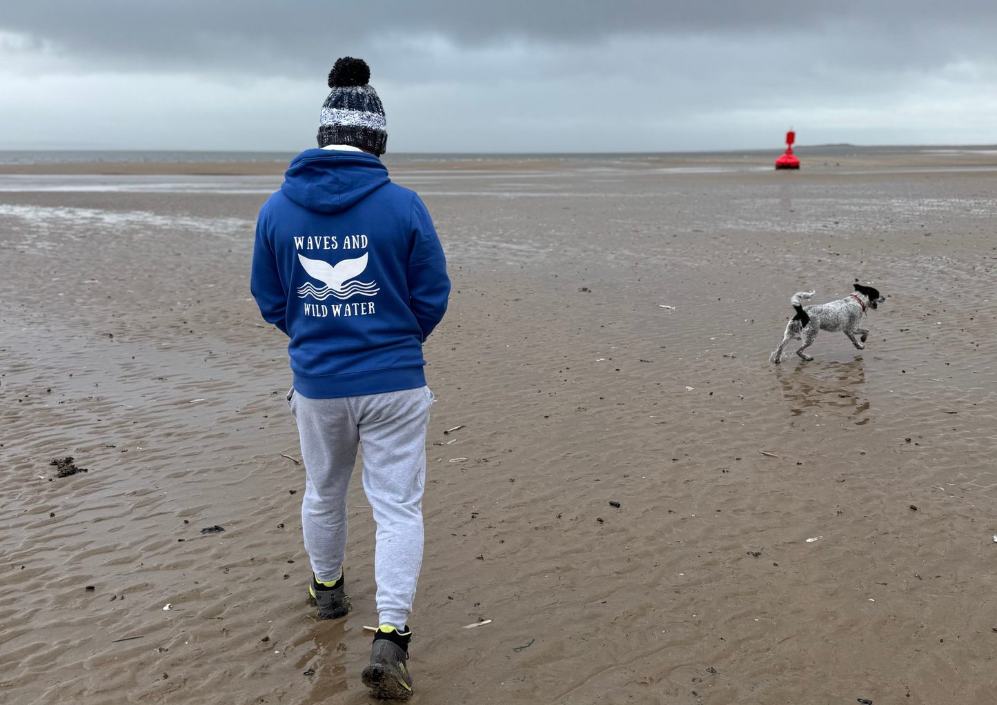 A man wearing a blue Waves & Wild Water hoodie on a beach. Printed by Pathfinder Printworks in South Wales.